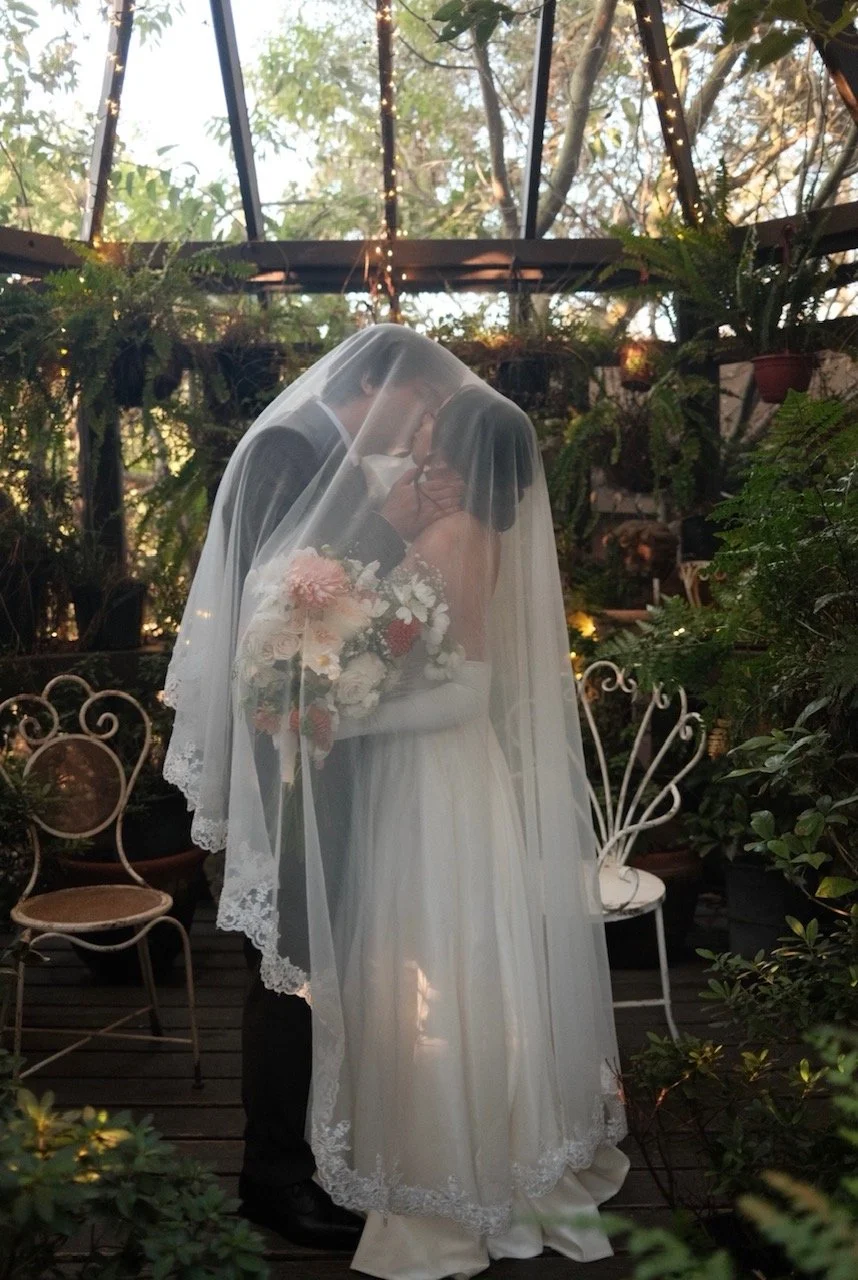 A bride and groom kiss under a veil on a wedding day, surrounded by greenery and decorated with string lights.