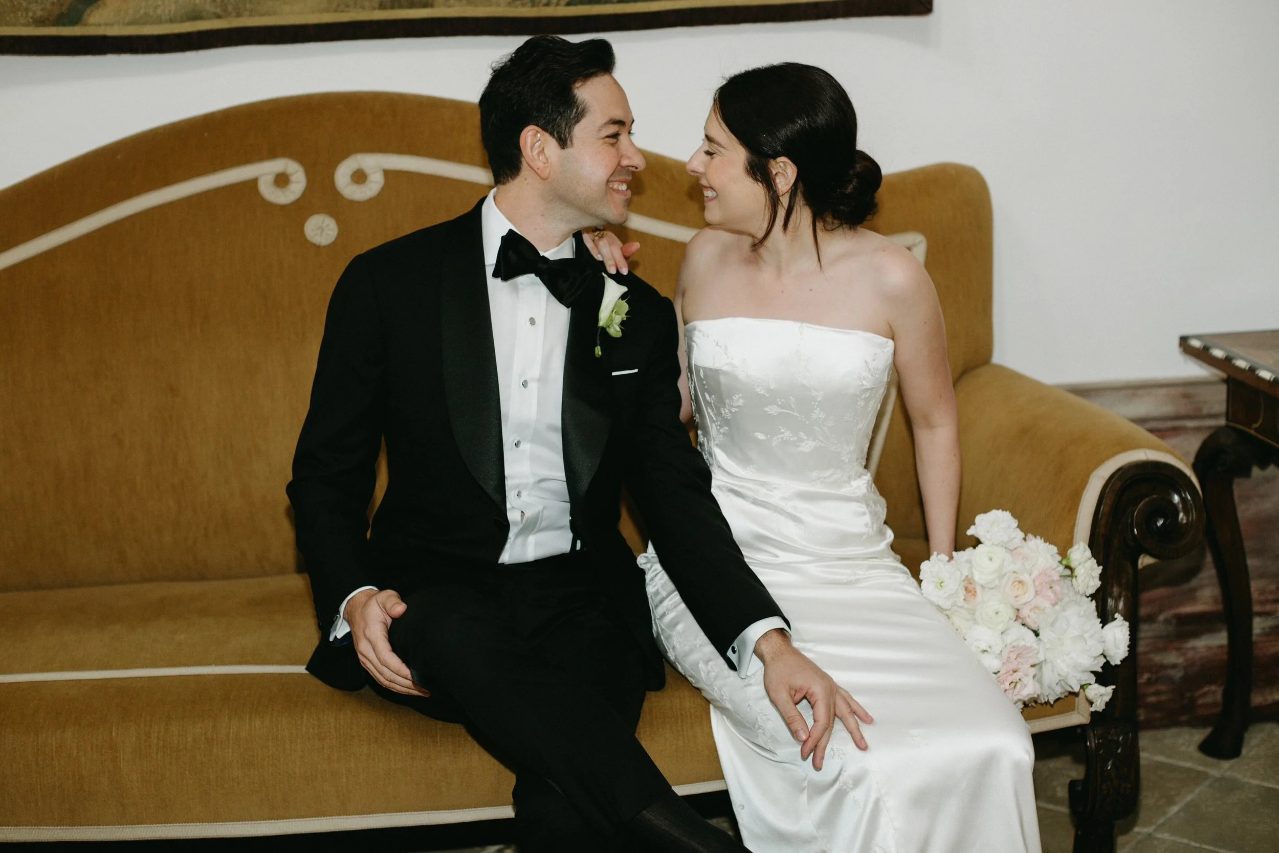 A bride and groom sitting together on a vintage sofa, smiling at each other. The groom is in a black tuxedo and the bride is in a white strapless wedding dress, holding a bouquet of white and pink flowers.