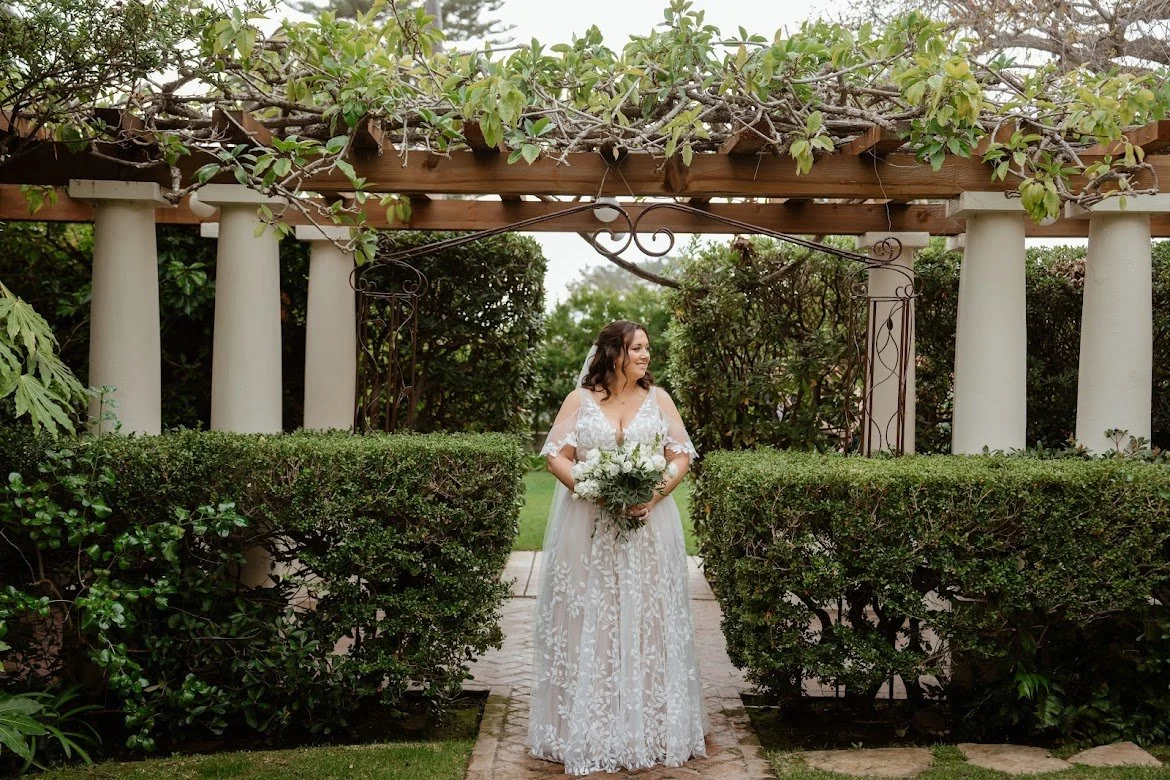 Bride in a white lace wedding dress standing in a garden with bushes, holding a bouquet of white flowers.