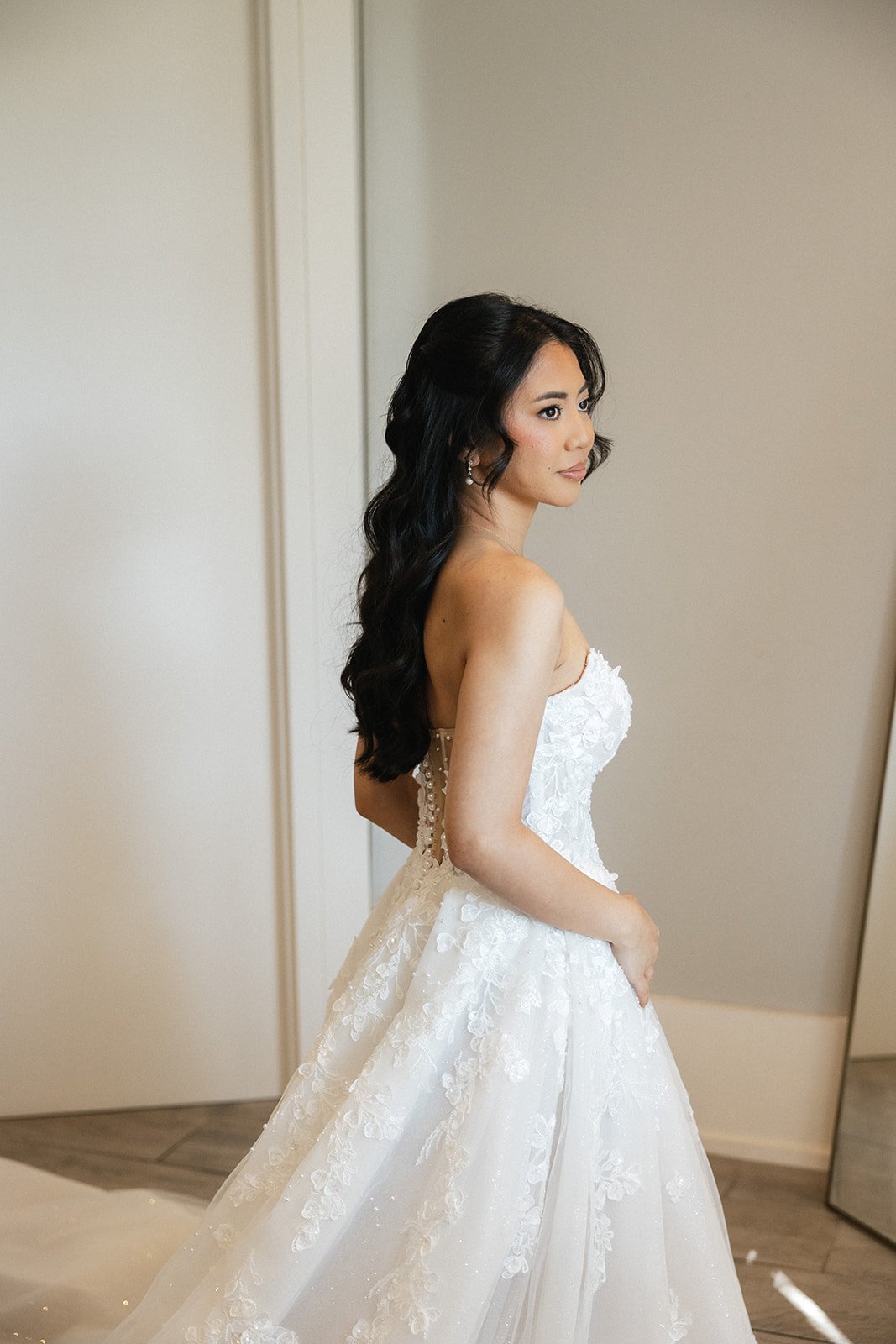 A woman in a white wedding dress with long dark hair standing in a room with a neutral-colored wall.