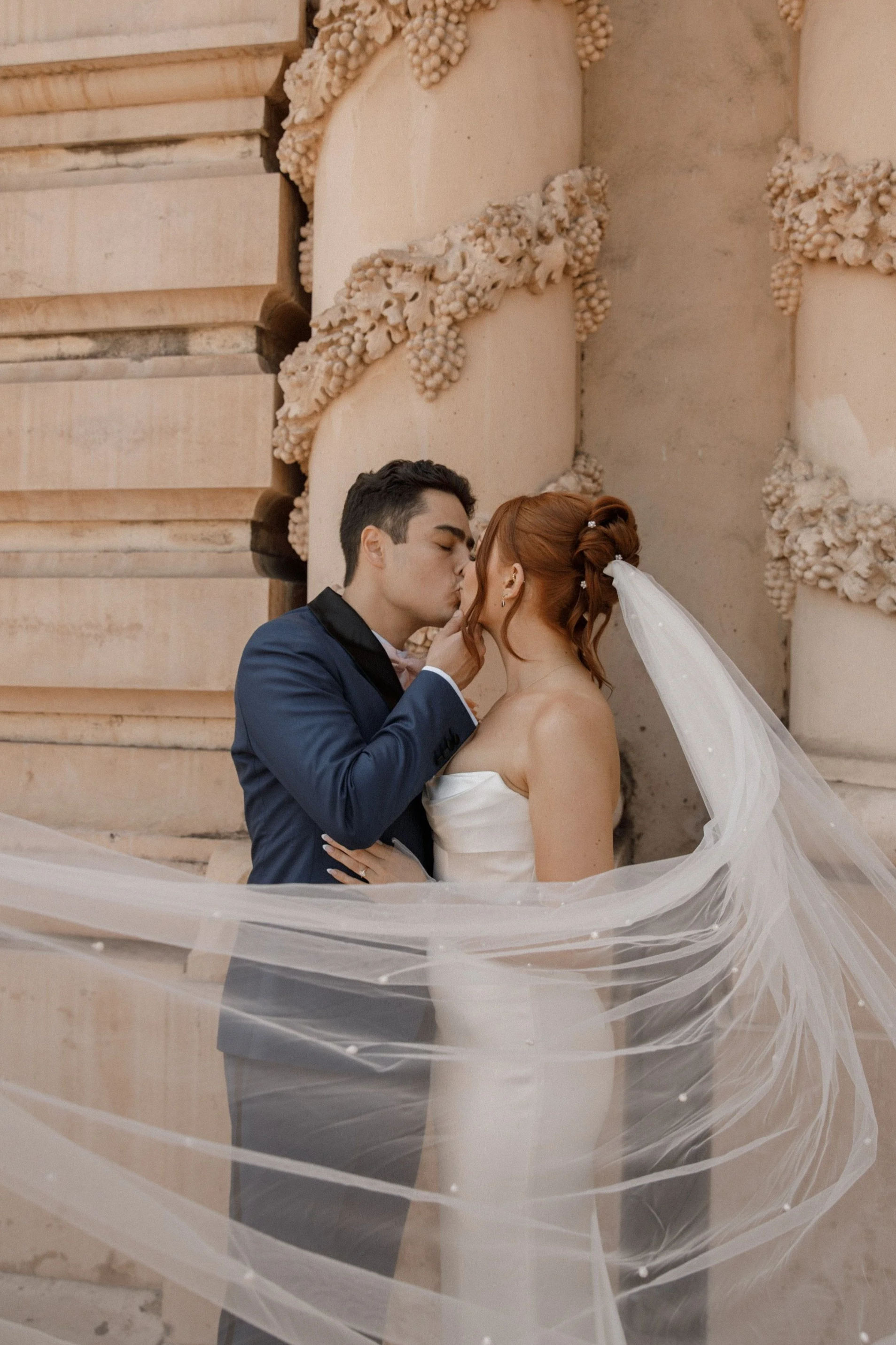 A bride and groom sharing a kiss, with the groom touching the bride's face, in front of ornate stone columns decorated with grapevine motifs, during their wedding photoshoot.