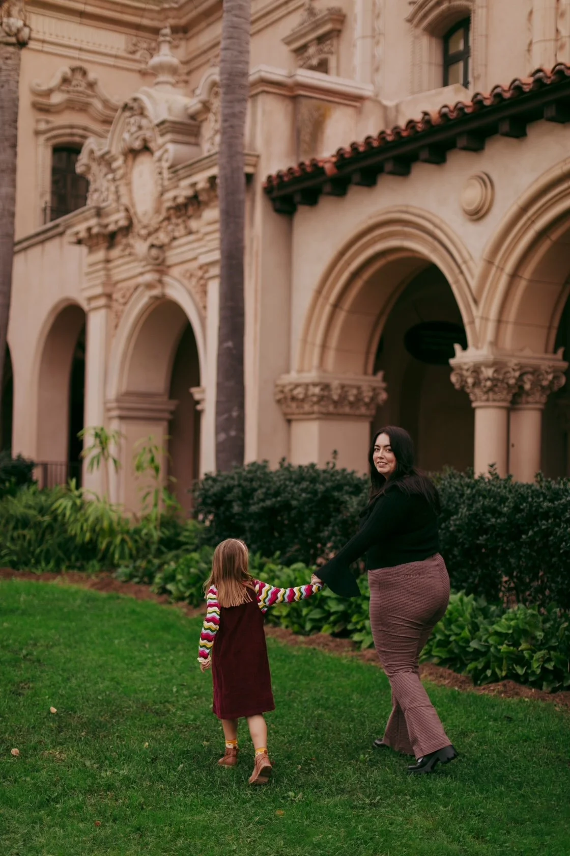 A woman and a young girl holding hands with a historic building with arches in the background. The woman has dark hair and is wearing a black top and brown pants, while the girl has red hair and is wearing a colorful striped shirt with a dark red dress.
