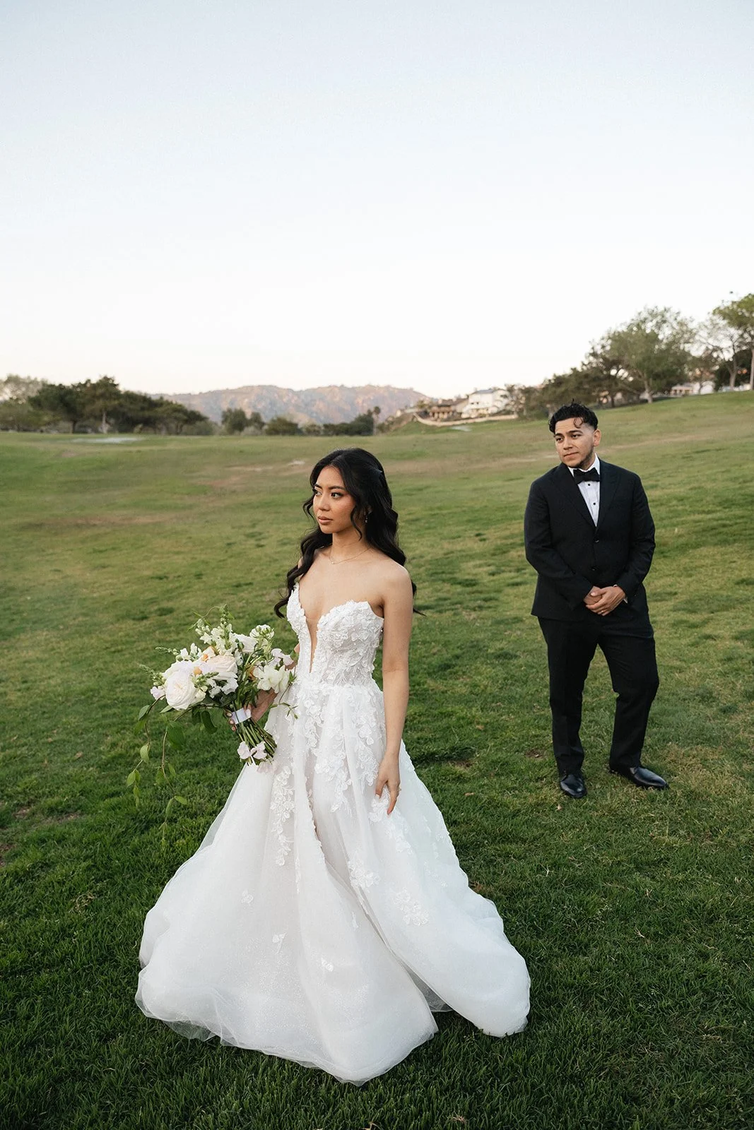 A bride in a white wedding dress holding a bouquet of flowers standing on a grassy field with a groom in a black tuxedo in the background.