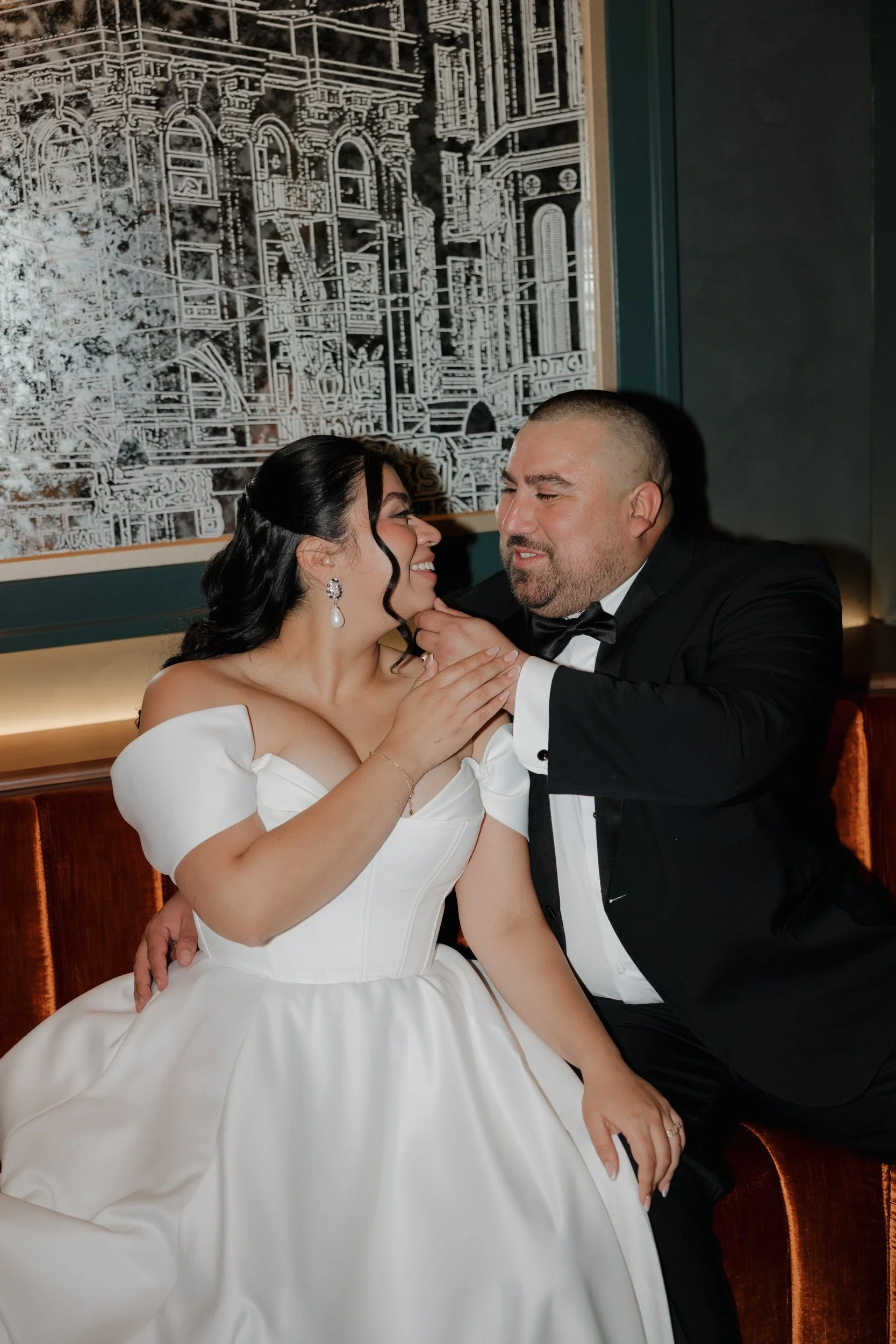 A bride in a white wedding gown and a groom in a tuxedo share an intimate moment, smiling and touching each other's faces while sitting on a bench against a decorative wall.