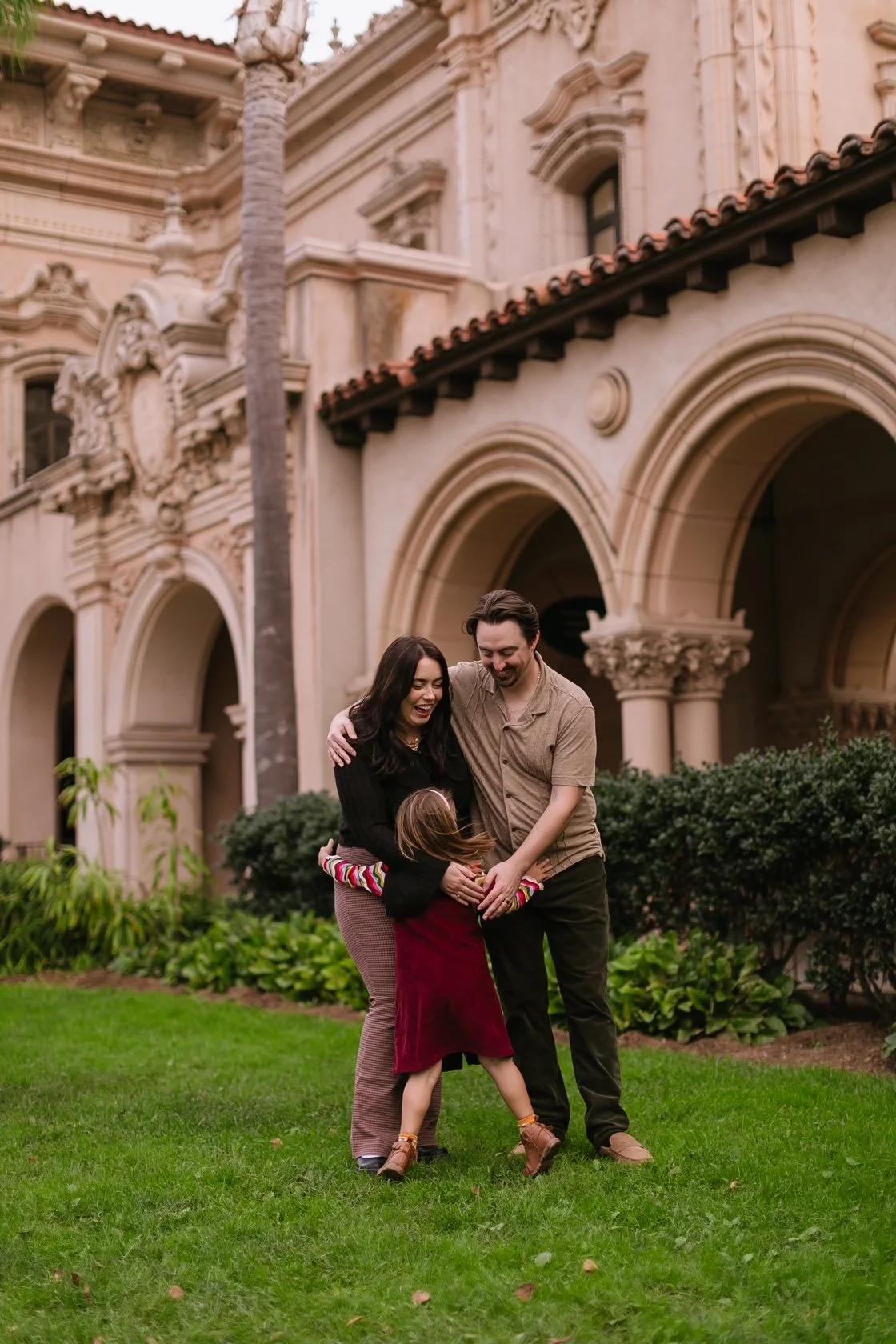 A happy family of three, a mother, father, and young daughter, playing and hugging on a lush green lawn in front of a historic European-style building with ornate architectural details.