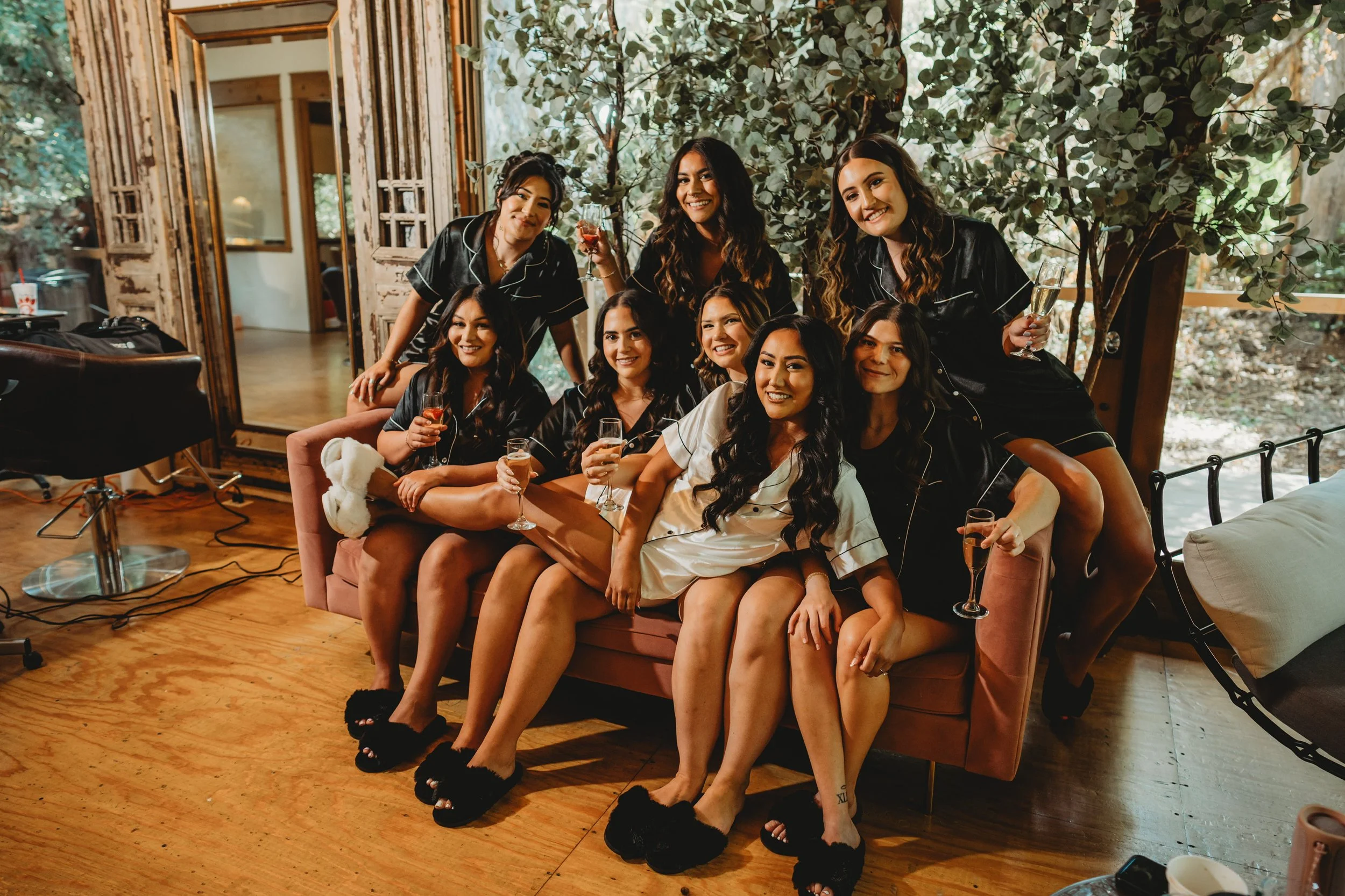 Group of women in pajamas sitting and standing on a pink sofa, celebrating with champagne and wine glasses in a cozy, decorated room with wooden floors and a large window with outdoor greenery.