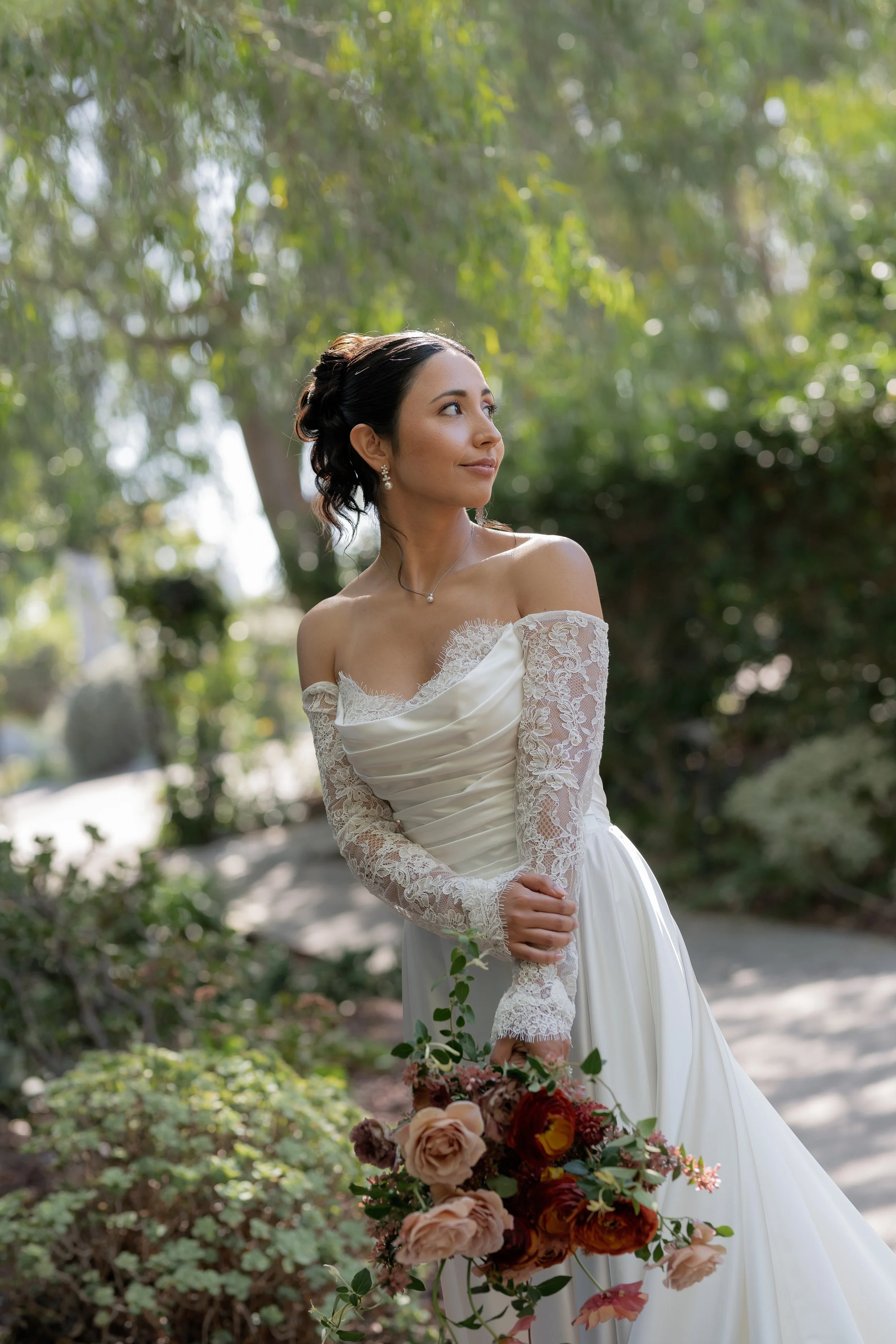 A bride standing outdoors in a white wedding dress with lace sleeves, holding a bouquet of pink and red flowers, surrounded by greenery and sunlight.