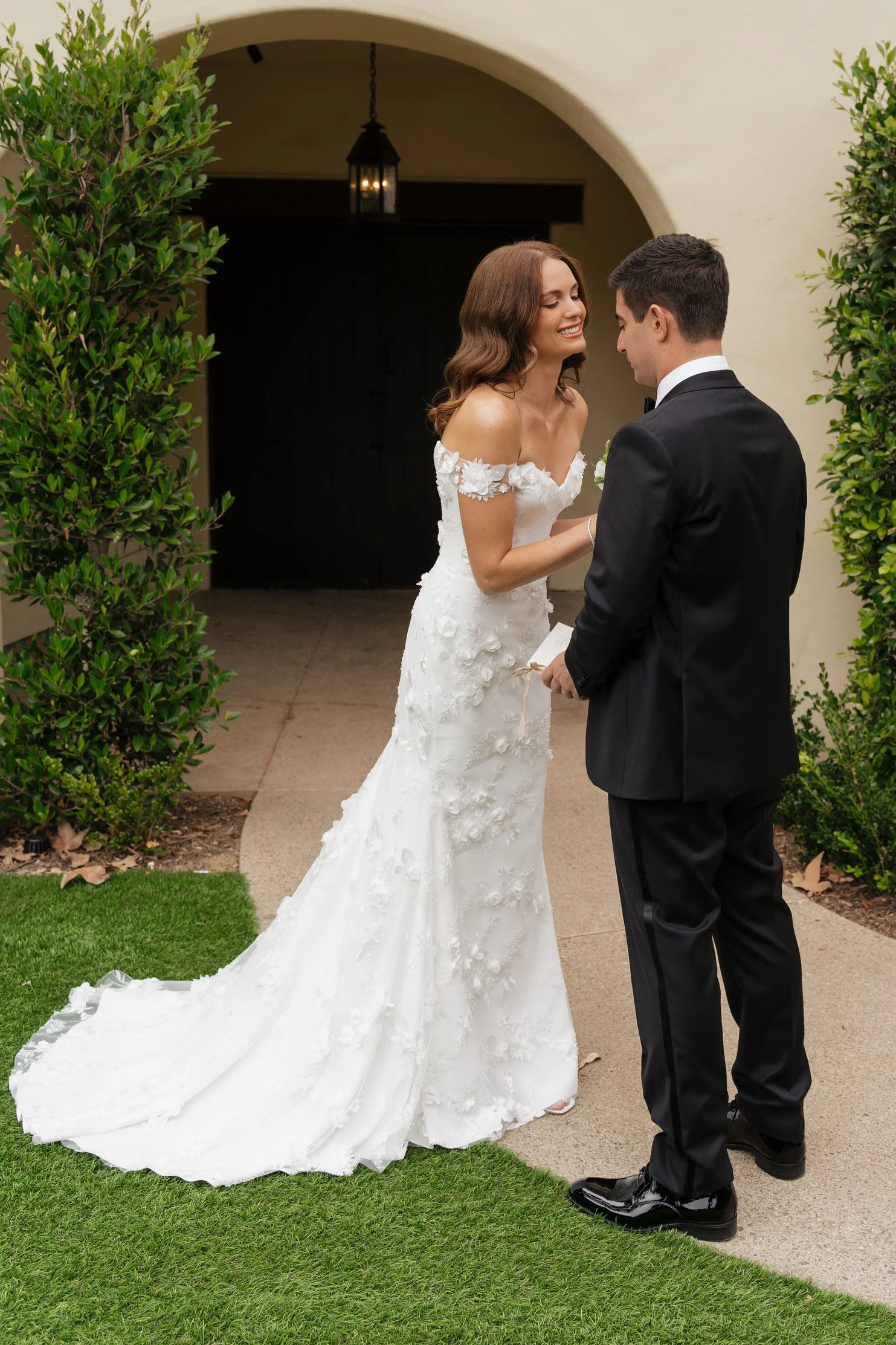 A bride and groom share a tender moment outside a building with an archway. The bride wears a white off-shoulder wedding gown with floral details, and the groom wears a black tuxedo. They are smiling and holding hands, standing on a grassy area with 