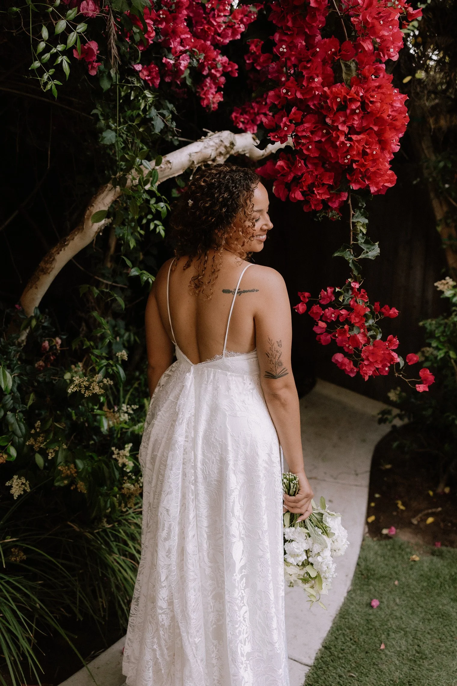 A woman in a white dress holding a bouquet of white flowers, standing in front of vibrant pink and red bougainvillea flowers and greenery, facing away with her head turned slightly to the side.