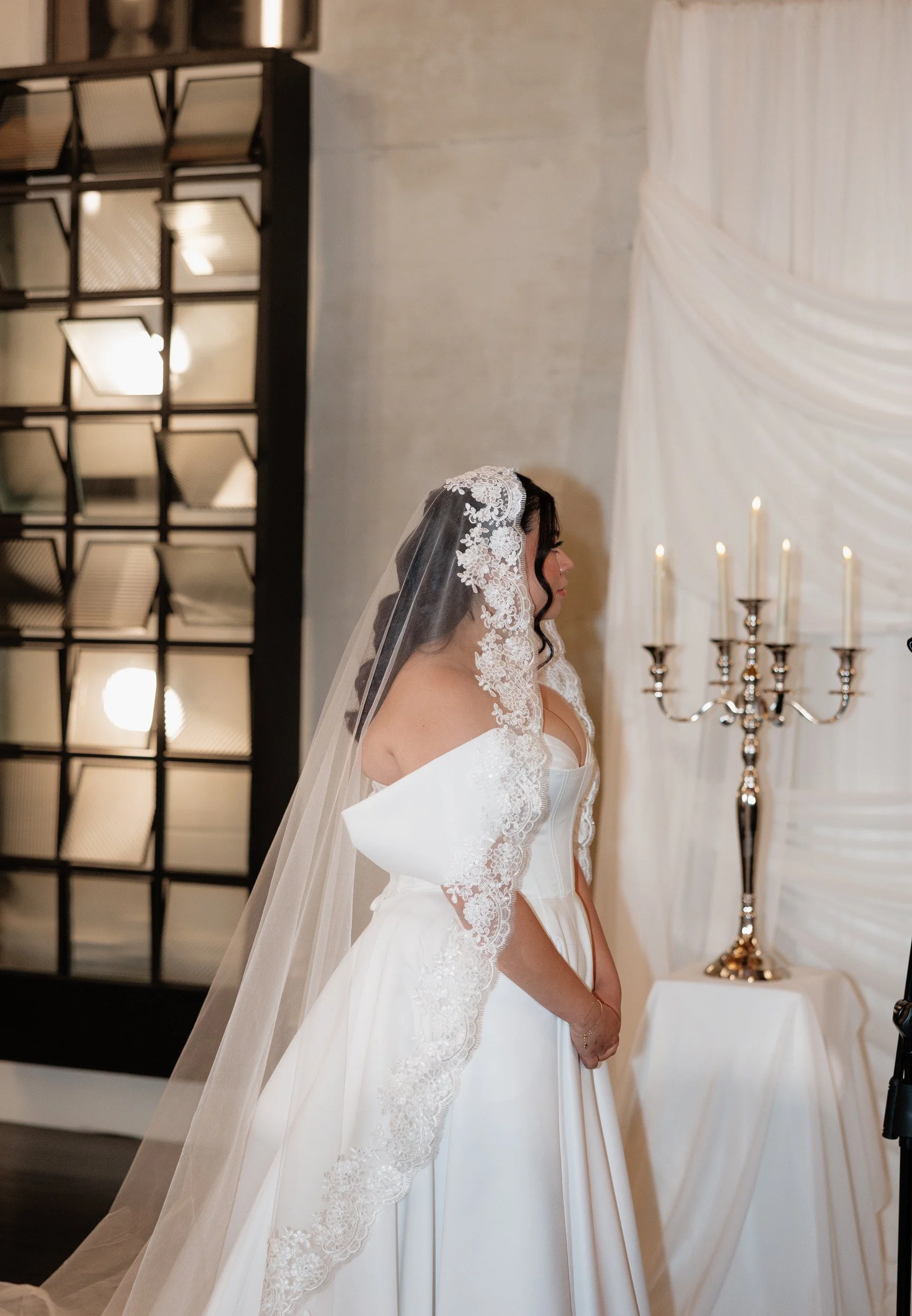 A bride in a white wedding dress and lace veil stands by a candelabra with lit candles during her wedding ceremony.