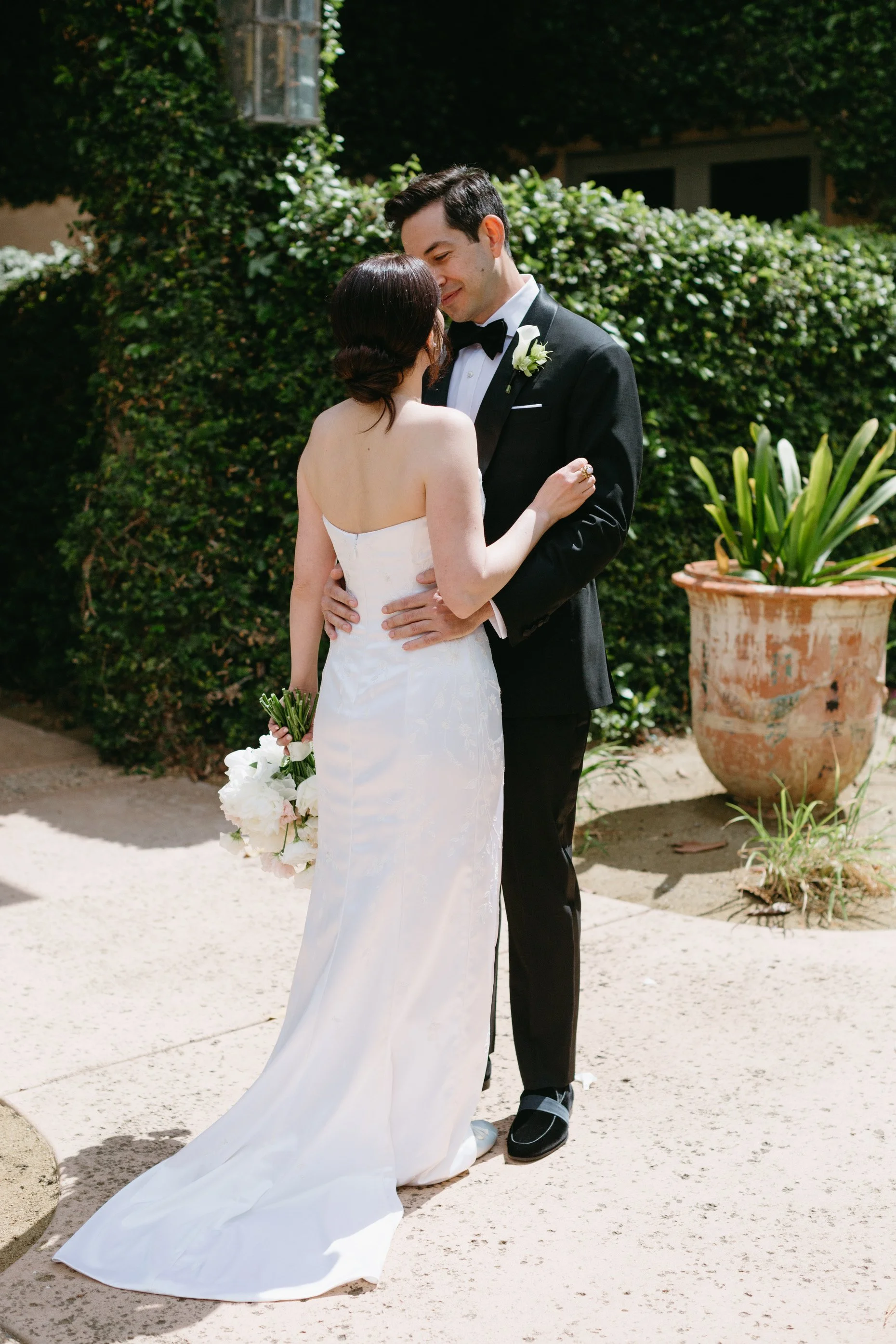 A bride and groom sharing a tender moment outdoors during their wedding, with the bride holding a bouquet of white and pink flowers, and the groom in a black tuxedo with a white boutonniere, standing on a paved pathway with lush greenery and potted p