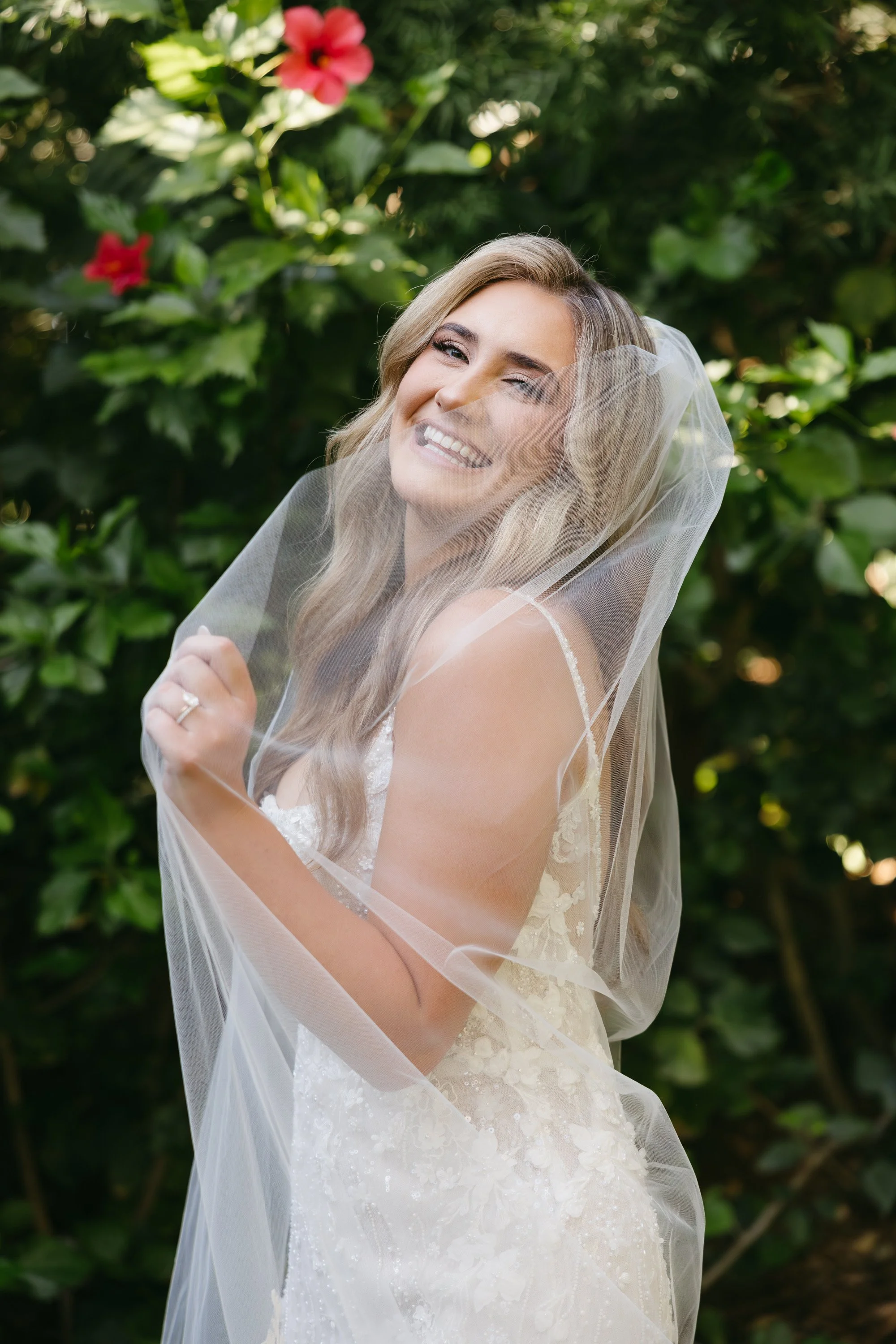 Bride with long blonde hair winks, smiling, wearing a white lace wedding dress and veil, outdoors with green foliage and pink flowers in background.