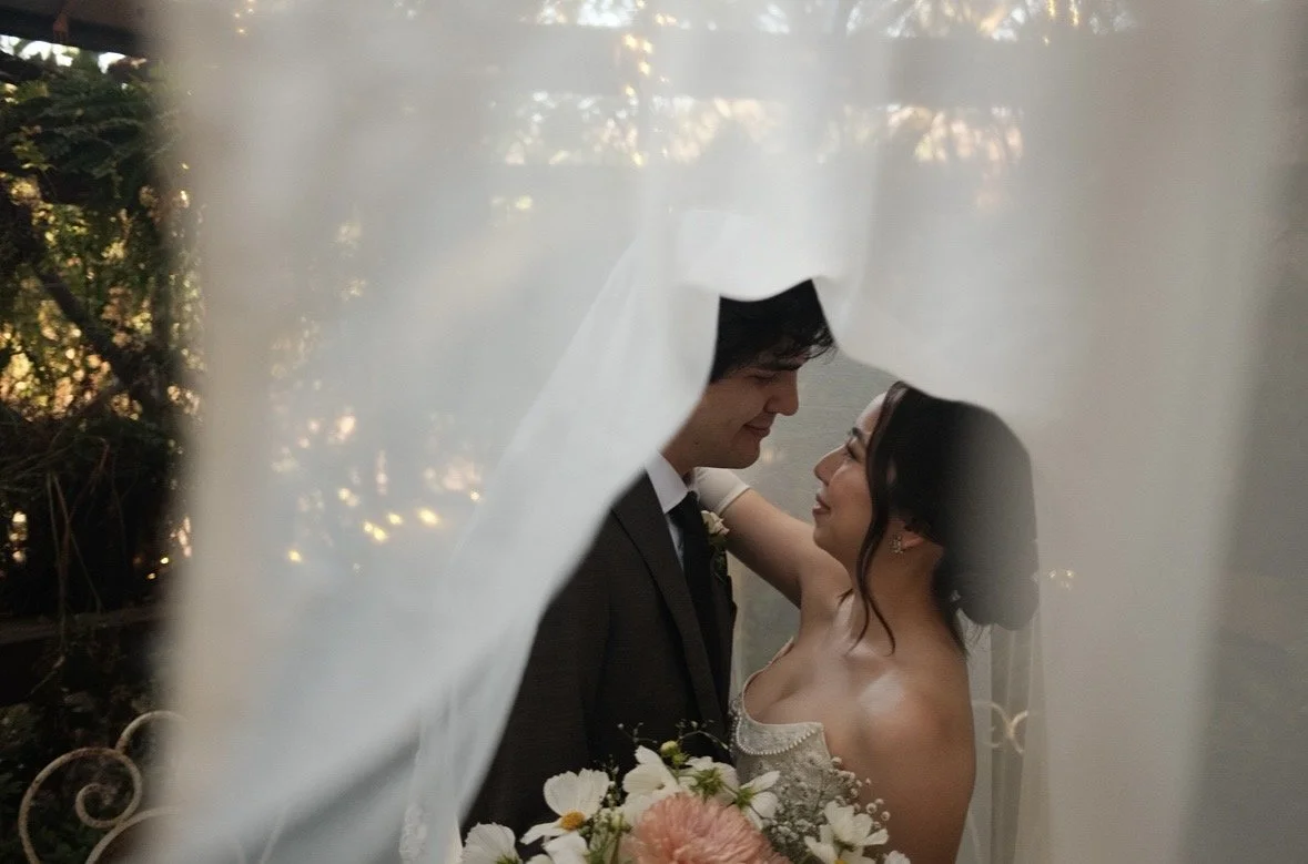 A bride and groom look at each other through a veil during their wedding, with the bride holding a floral bouquet.