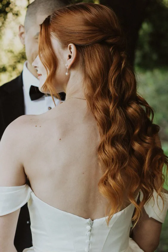 Close-up of a bride with long, wavy red hair wearing a white dress and pearl earrings, standing close to a groom in a tuxedo with a bow tie.