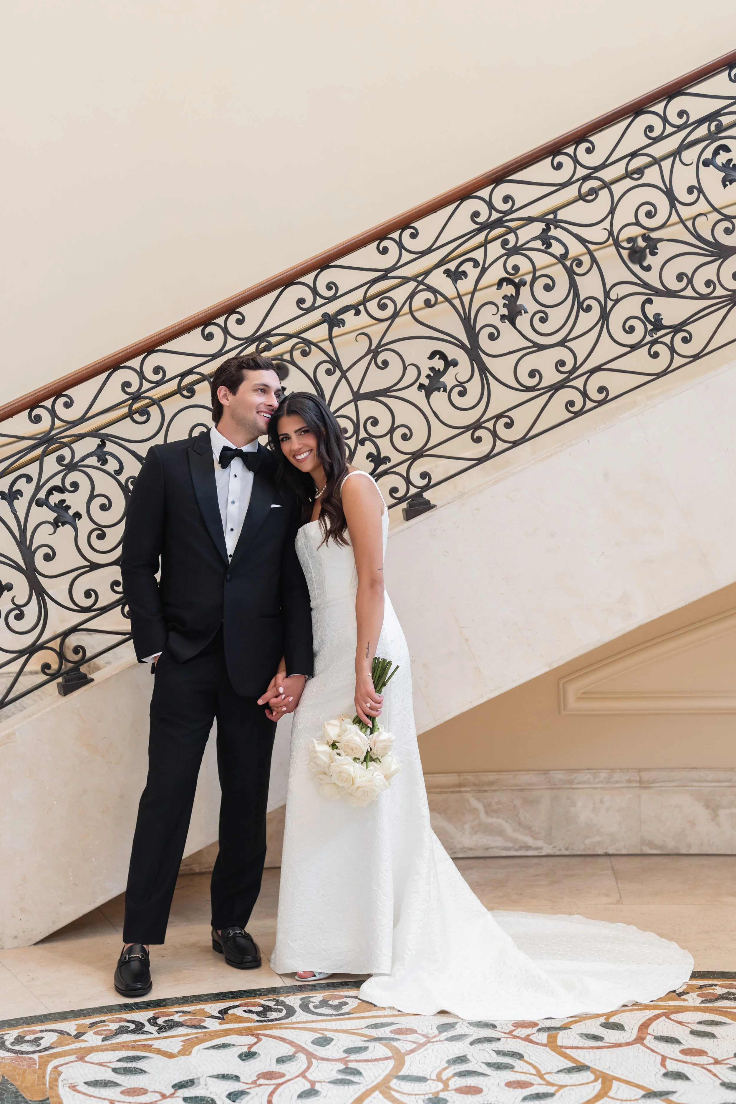 A newlywed couple dressed in wedding attire, standing indoors on a marble floor in front of a staircase with ornate wrought iron railing, smiling and holding hands, the woman holding a bouquet of white roses.