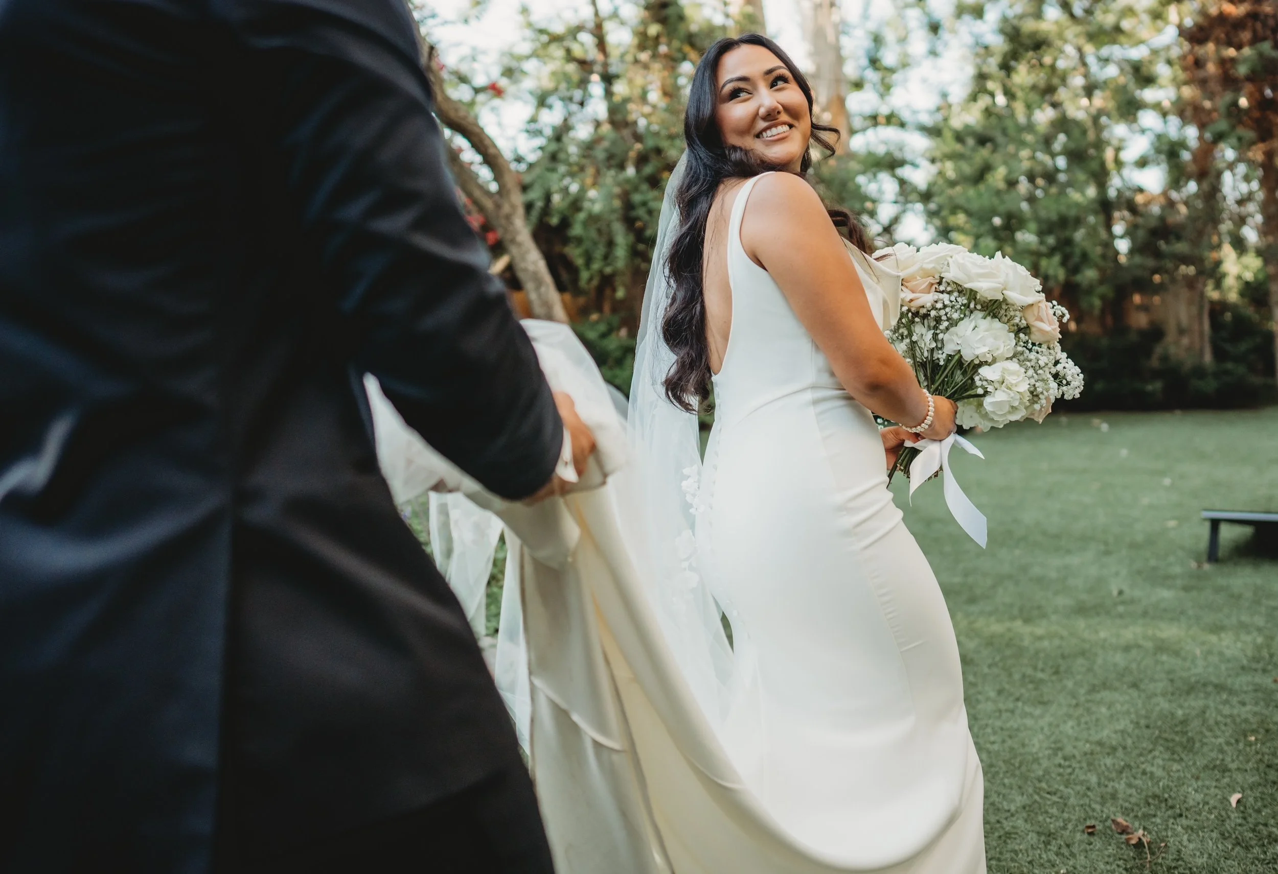 A smiling bride in a white wedding dress holding a bouquet of white flowers, walking outdoors on grass with a man in a black suit holding her hand.