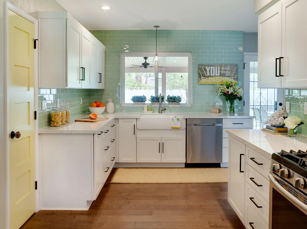 Coastal cottage kitchen remodel featuring seafoam green subway tile, white shaker cabinetry, a farmhouse sink, stainless steel appliances, and a cheerful yellow pantry door.