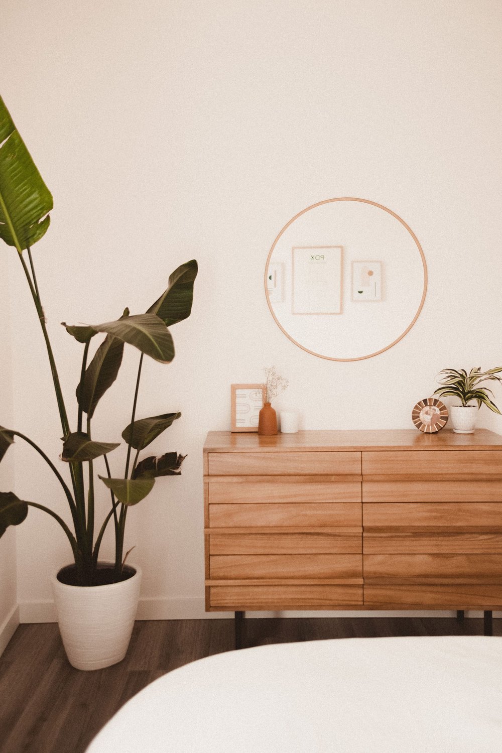  Warm wood toned dresser with minimalist decorative accents, round gold-framed mirror and large potted houseplant in a Portland home. 