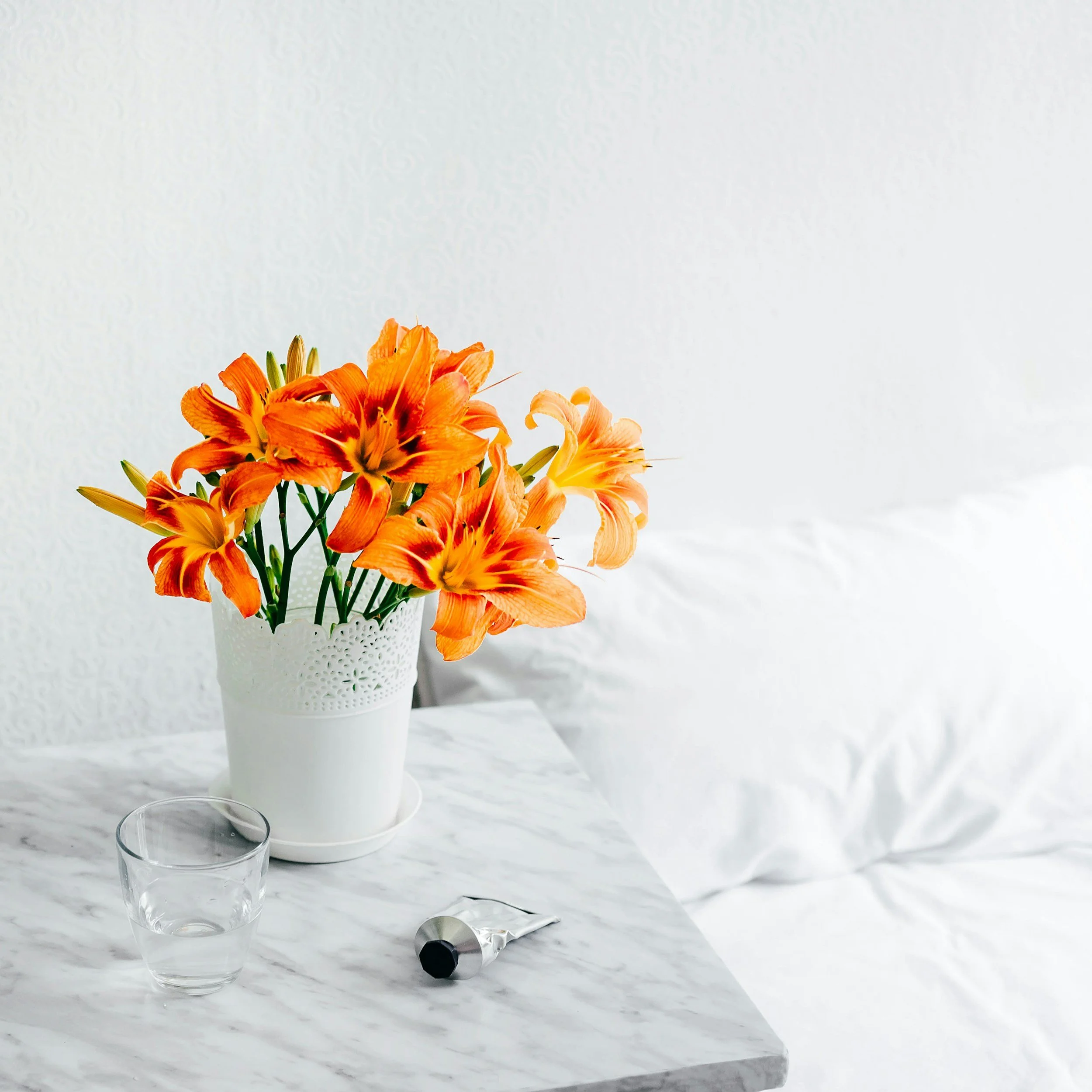 A bouquet of orange lilies in a white decorative vase on a white marble surface with a glass and a tube of cream or lotion.