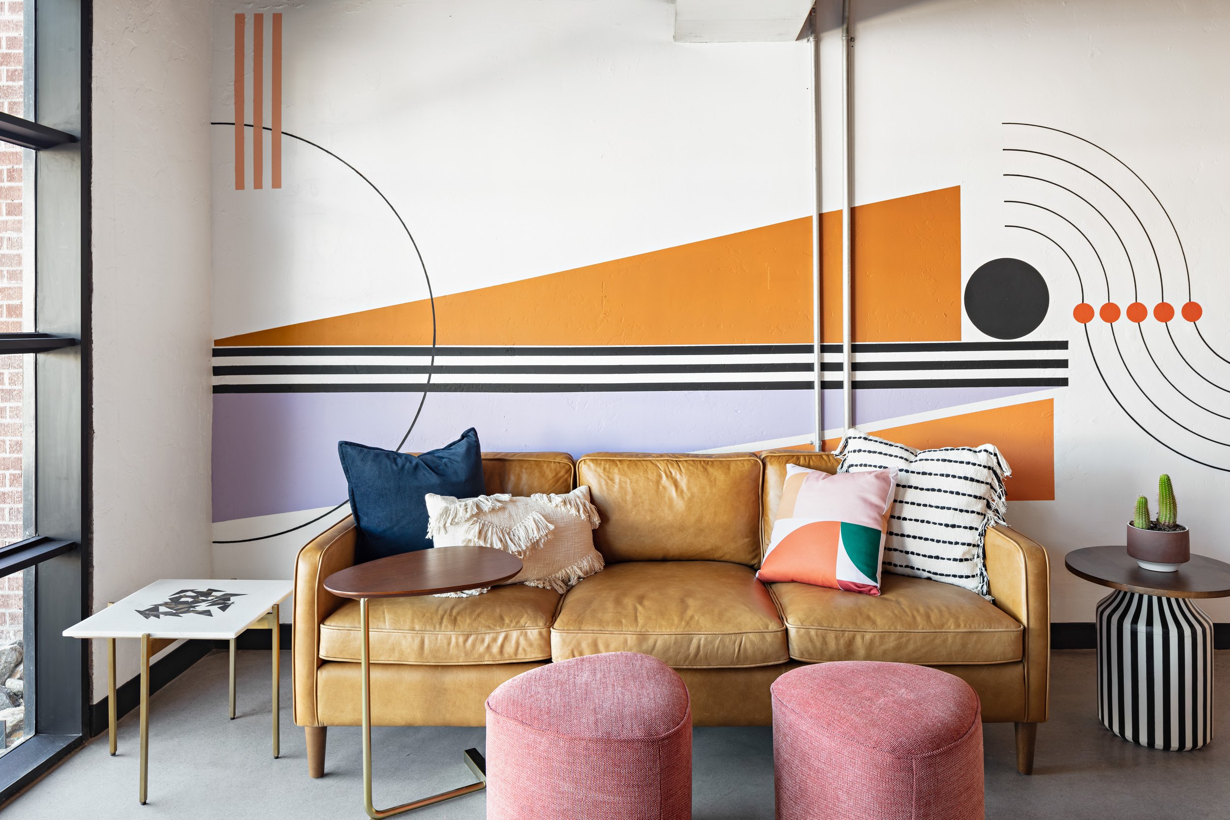 A vintage looking leather sofa, eclectic side tables, soft pink ottomans and bright throw pillows sit in front of a wall mural in a mid century modern office in Scottsdale.