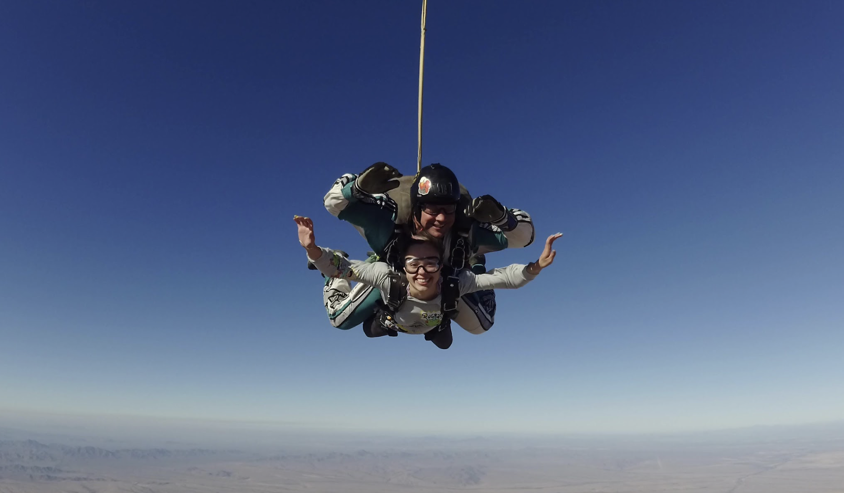 Two skydivers in mid-air, smiling and free-falling over a desert landscape with clear blue sky.