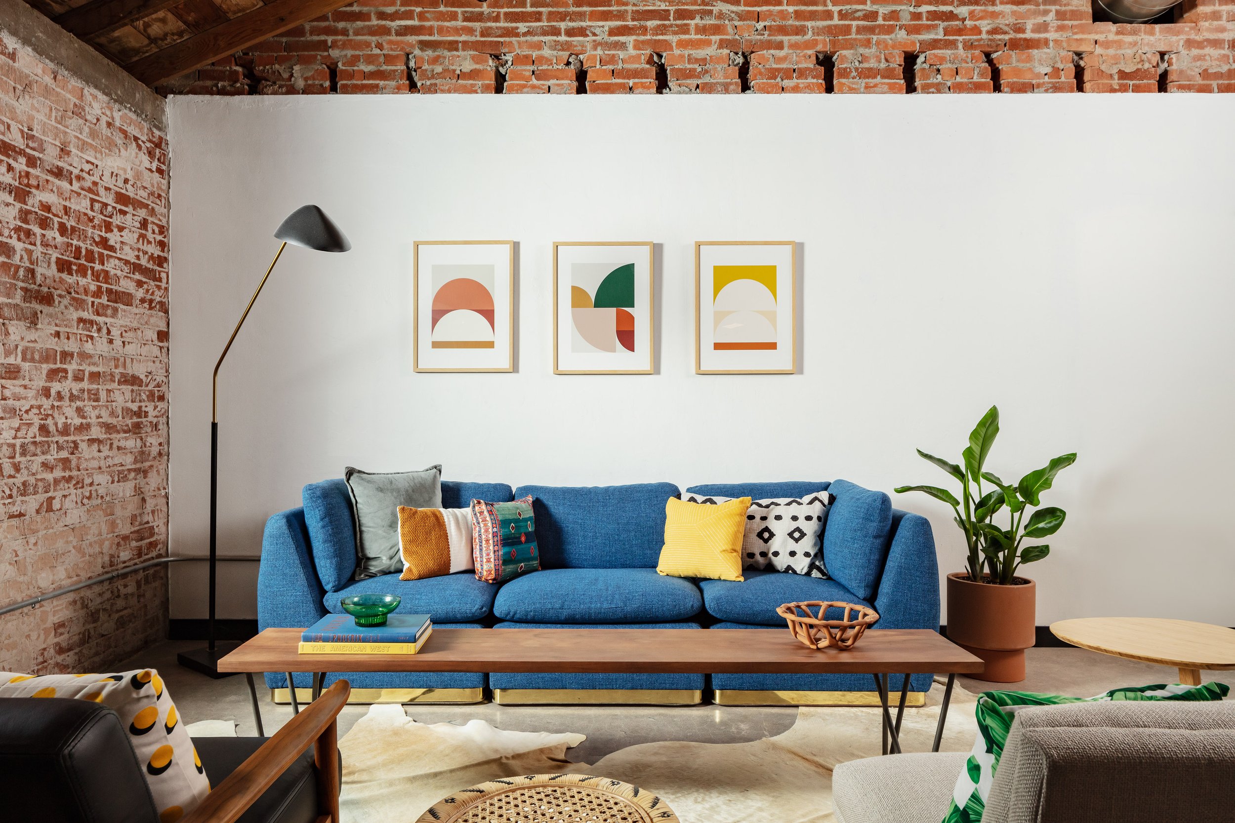 Mid century modern office with exposed brick and exposed ceilings, a modern blue sofa, industrial coffee table, colorful wall art and potted plants.
