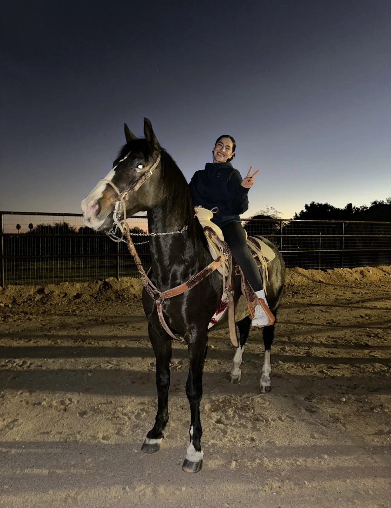 A person smiling and posing with a peace sign while riding a black horse at dusk, with a fenced outdoor riding area and trees in the background.