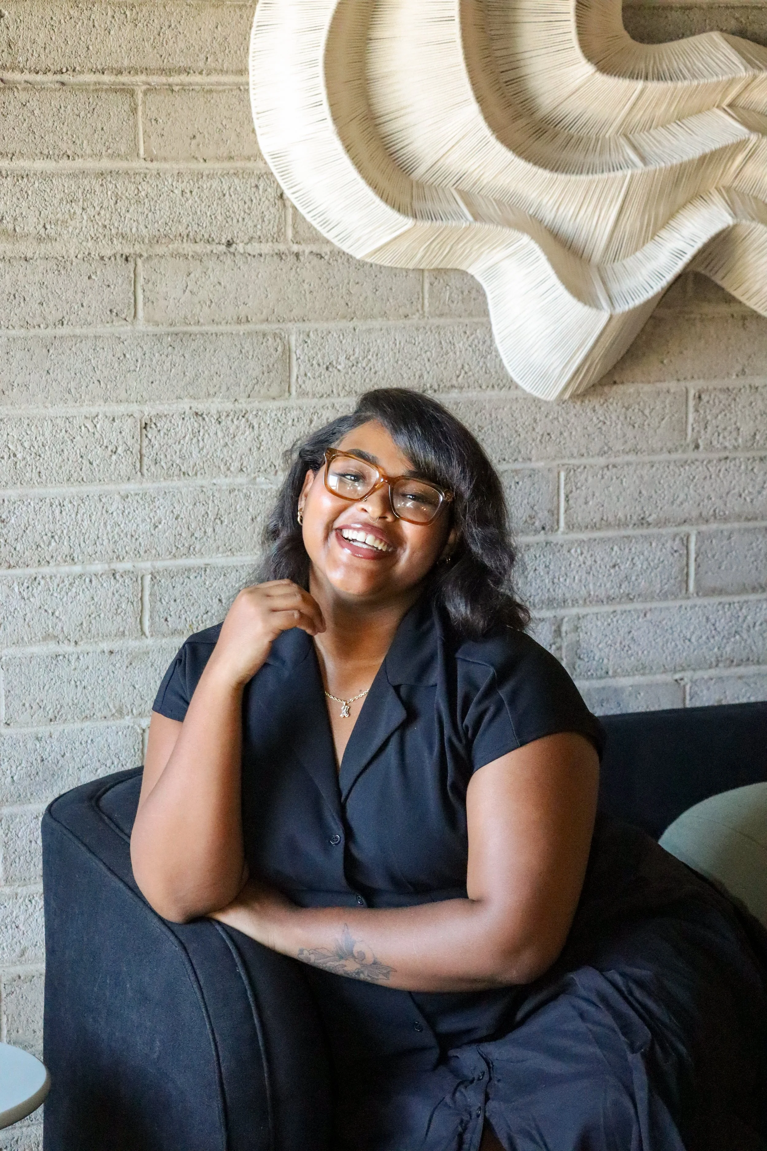 A smiling woman with glasses sitting on a black couch in front of a beige brick wall with a large, abstract wooden wall art.
