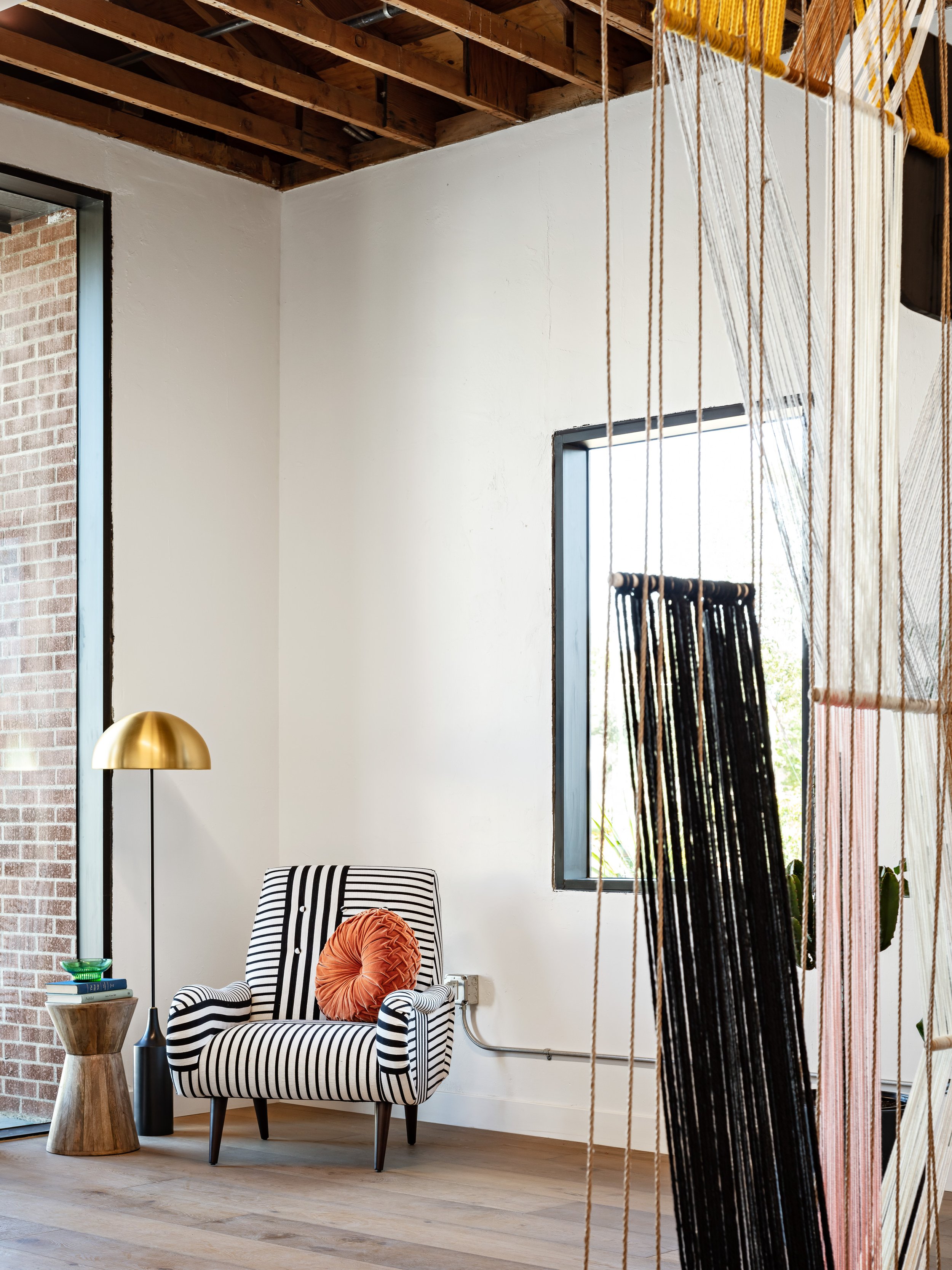 Custom art hangs from the exposed ceiling in a Scottsdale office space. A striped accent chair with colorful throw pillow and brass floor lamp sit in the entryway.