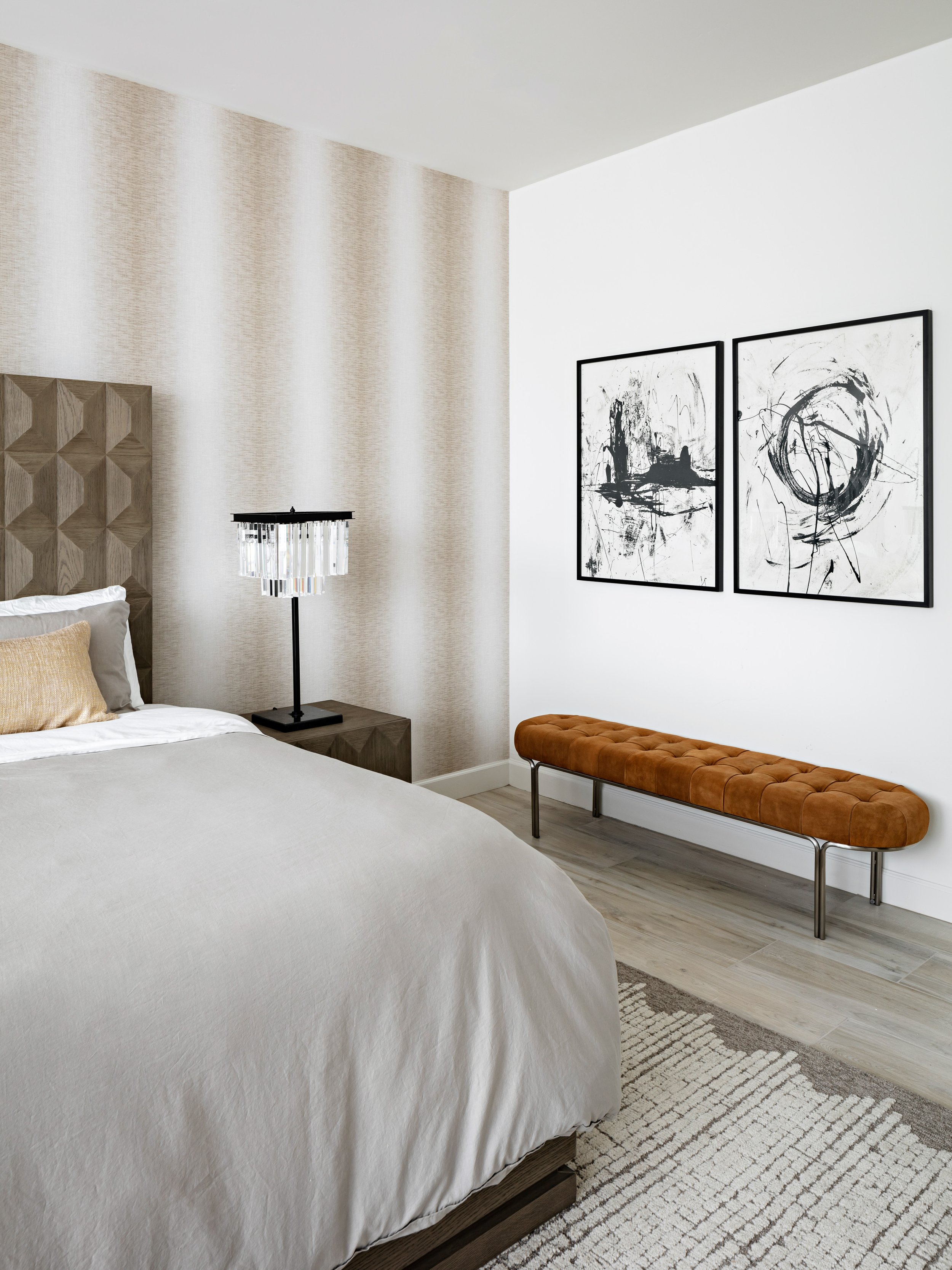 Monochromatic bedroom with textured neutral linens, area rug, headboard and wall paper. Black and white wall art hang above a leather bench.