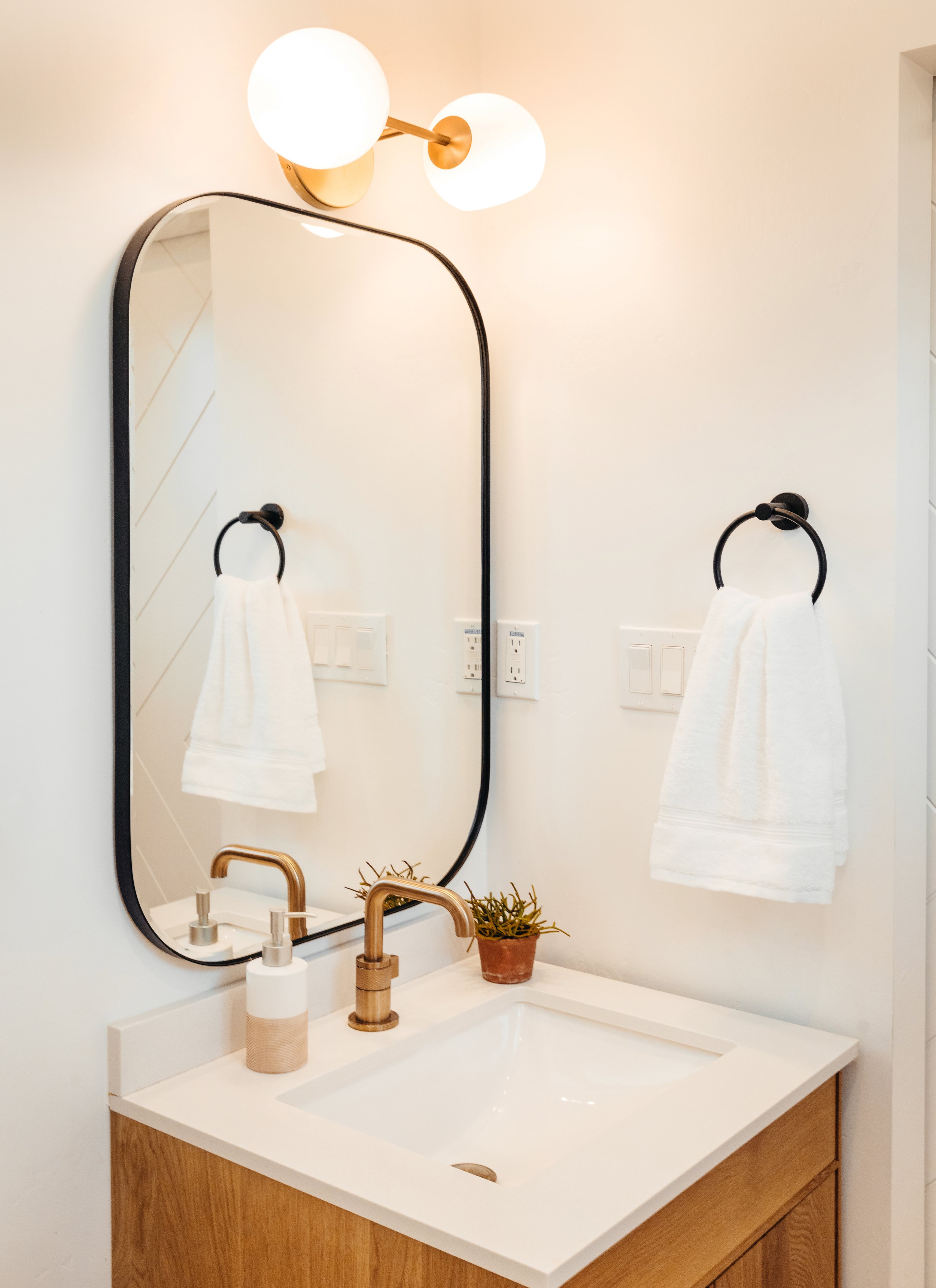 Rounded rectangle mirror in a polished bathroom with modern vanity, oak cabinets, and gold faucet.