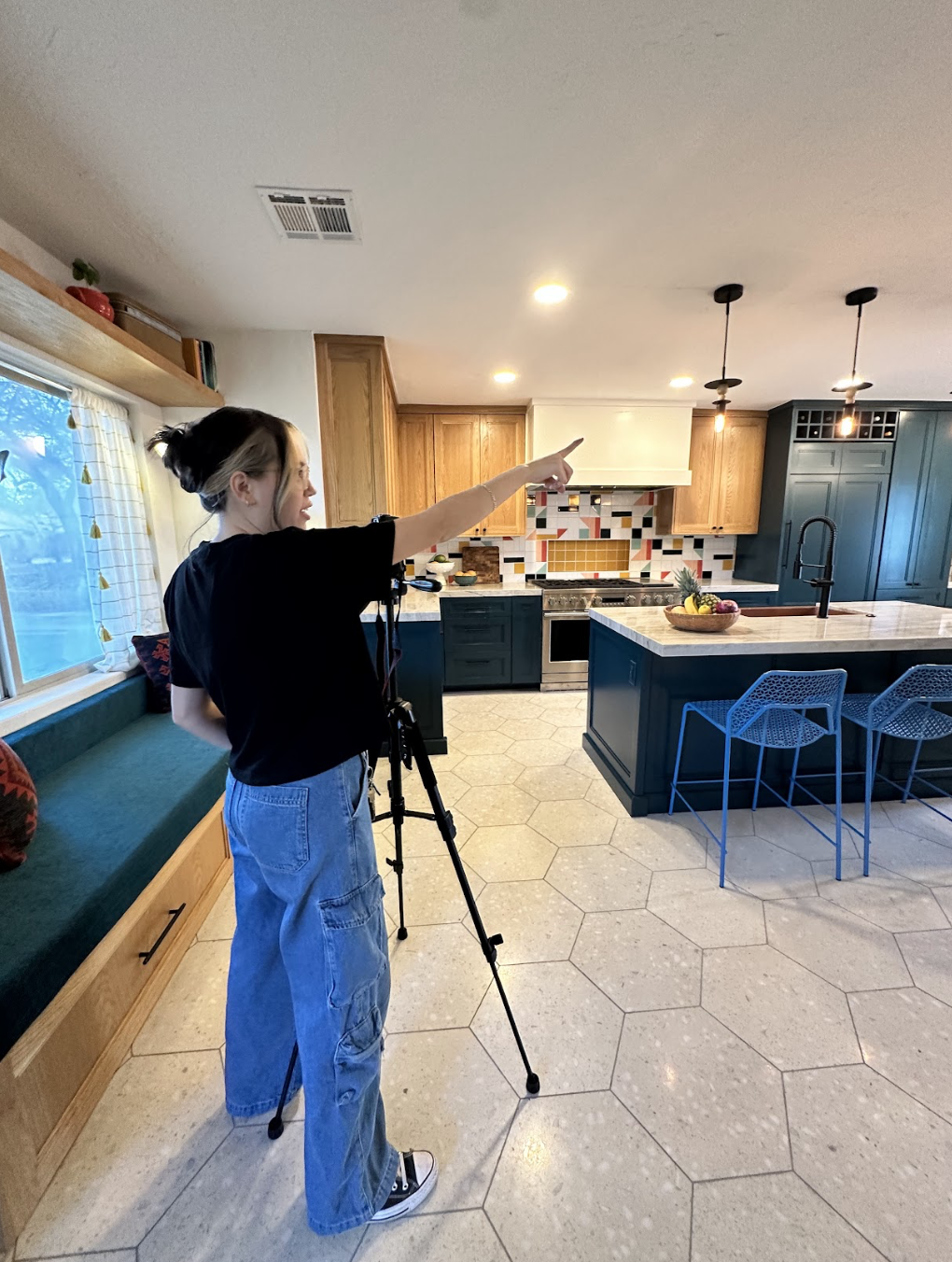 A woman in black shirt and baggy jeans is standing in a modern kitchen, pointing towards the ceiling while filming with a camera on a tripod. The kitchen has hexagonal tiled flooring, wooden cabinets, and a colorful tiled backsplash.