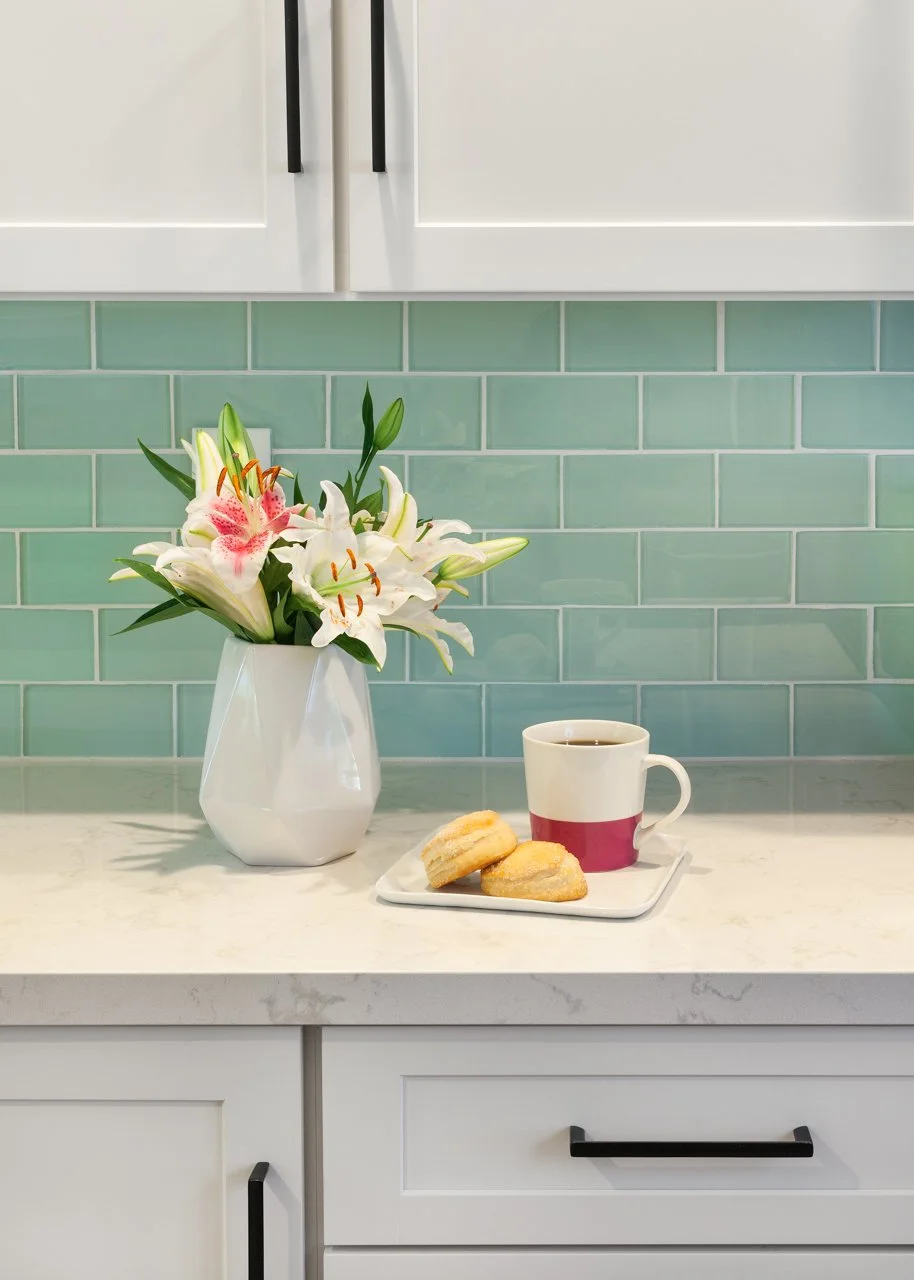 Close-up of glossy seafoam green subway tile backsplash, white shaker cabinets, and marble-look countertops with fresh lilies in the Cool Coastal Cottage kitchen.