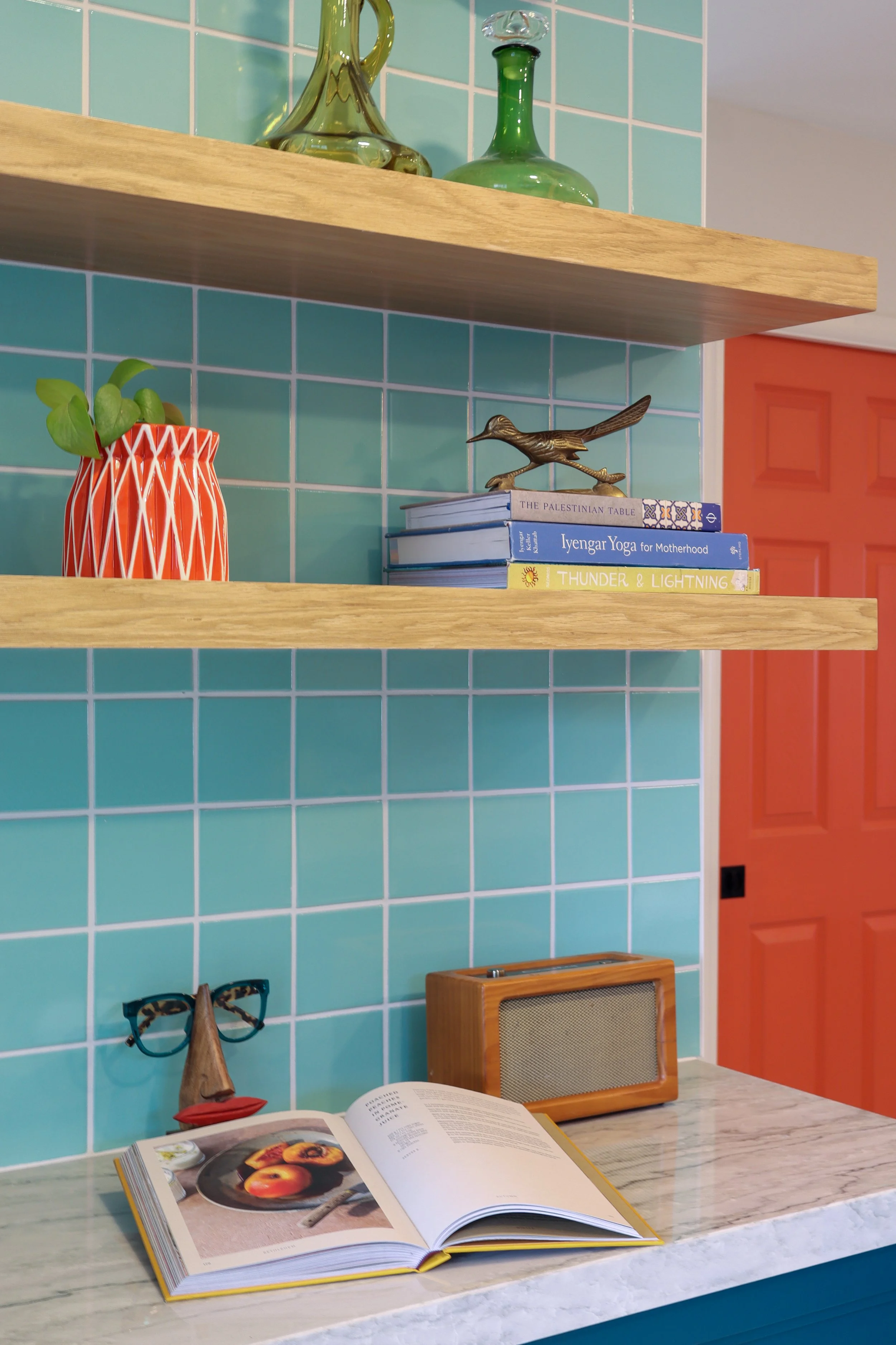Open cookbook with a photograph of peaches on a black plate on a marble countertop. In the background, colorful objects include a brown wooden radio, a pair of glasses with tortoiseshell frames, a small sculpture of a woman, a small photograph in a black frame, and a bookshelf with books and decorative items on pastel turquoise tiled walls with a mustard-colored door.