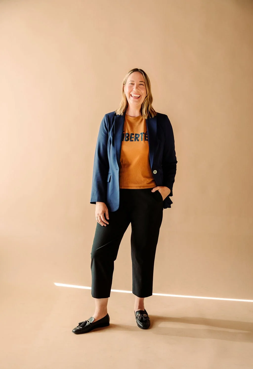 A woman smiling and laughing, wearing a navy blazer, an orange T-shirt, black pants, and loafers, standing against a plain beige wall.