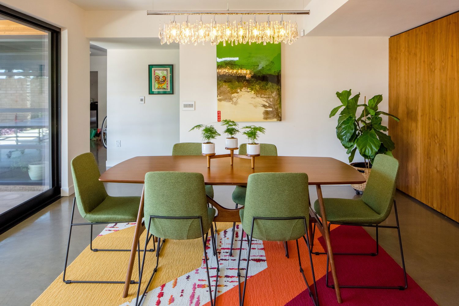 The vibrant dining area of this Tempe, Arizona remodel includes potted plants, colorful rug, and eclectic wall art. Vintage green chairs with backs wrap around a mid-century modern dining table.