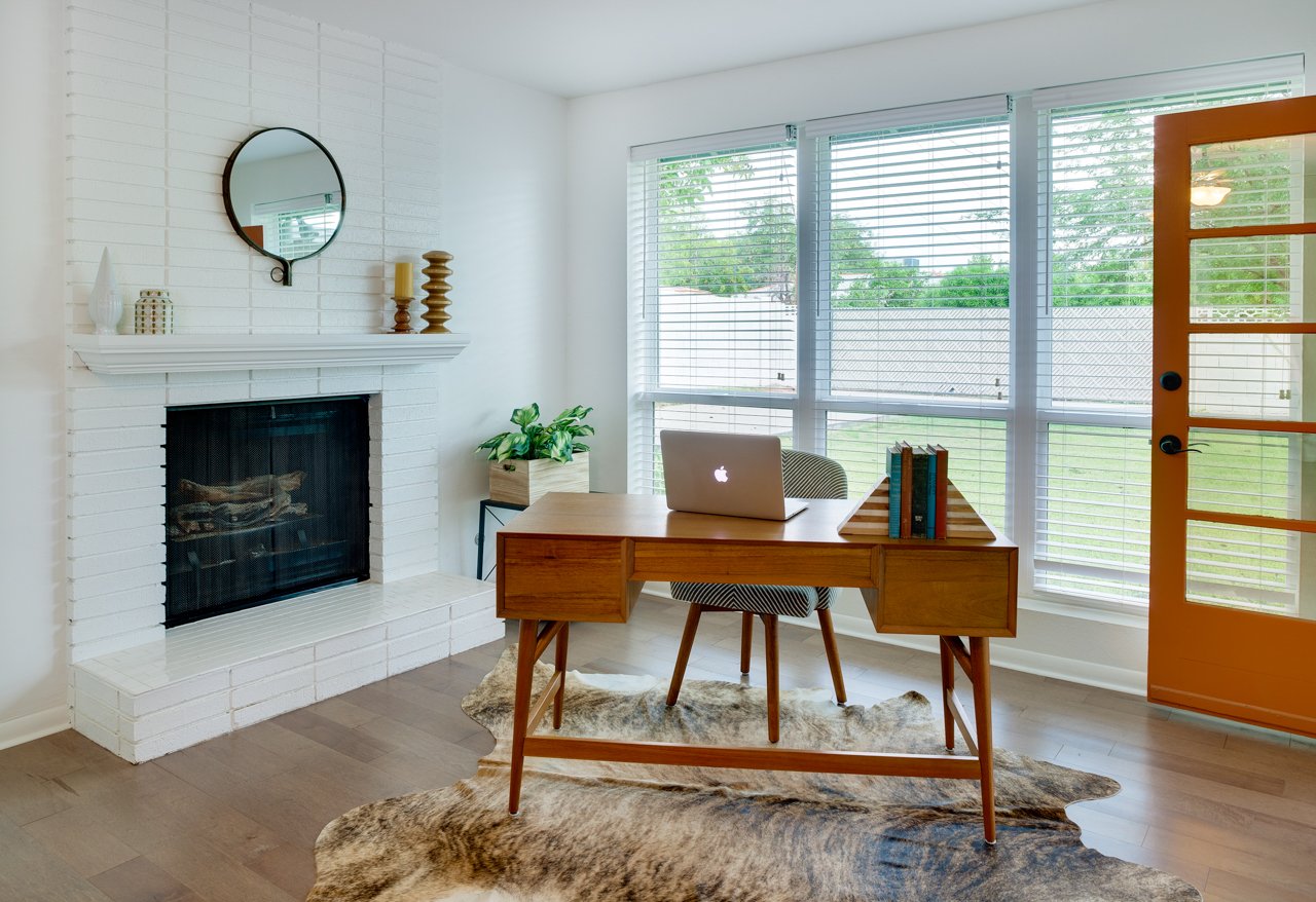 Light-filled coastal cottage home office featuring a midcentury modern wood desk, white painted brick fireplace, natural hide rug, and bright orange door.
