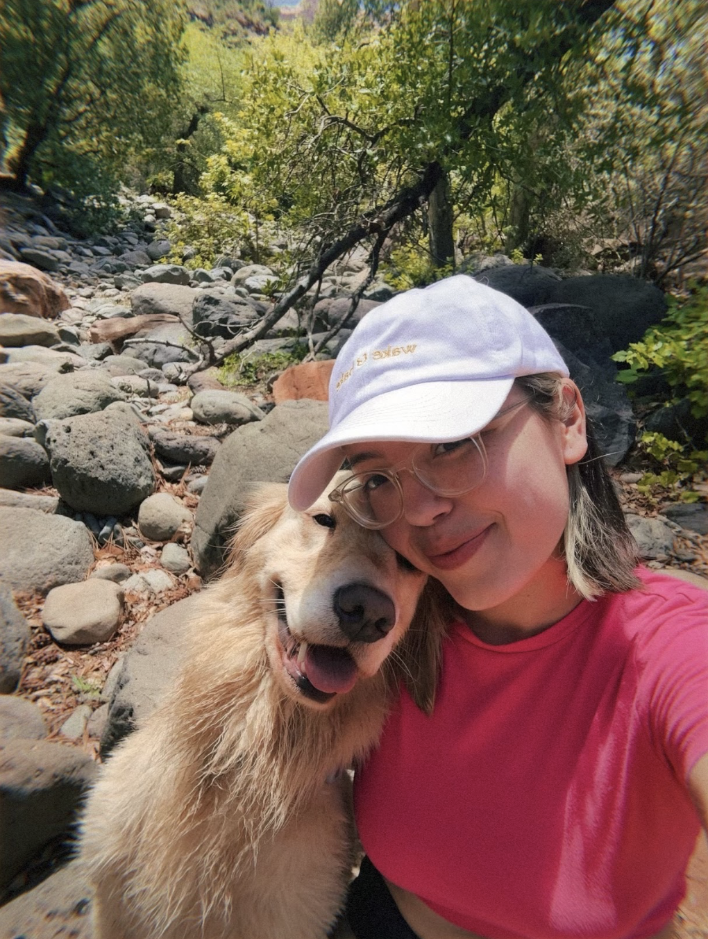A woman and a golden retriever dog taking a selfie together outdoors on a rocky trail surrounded by trees.