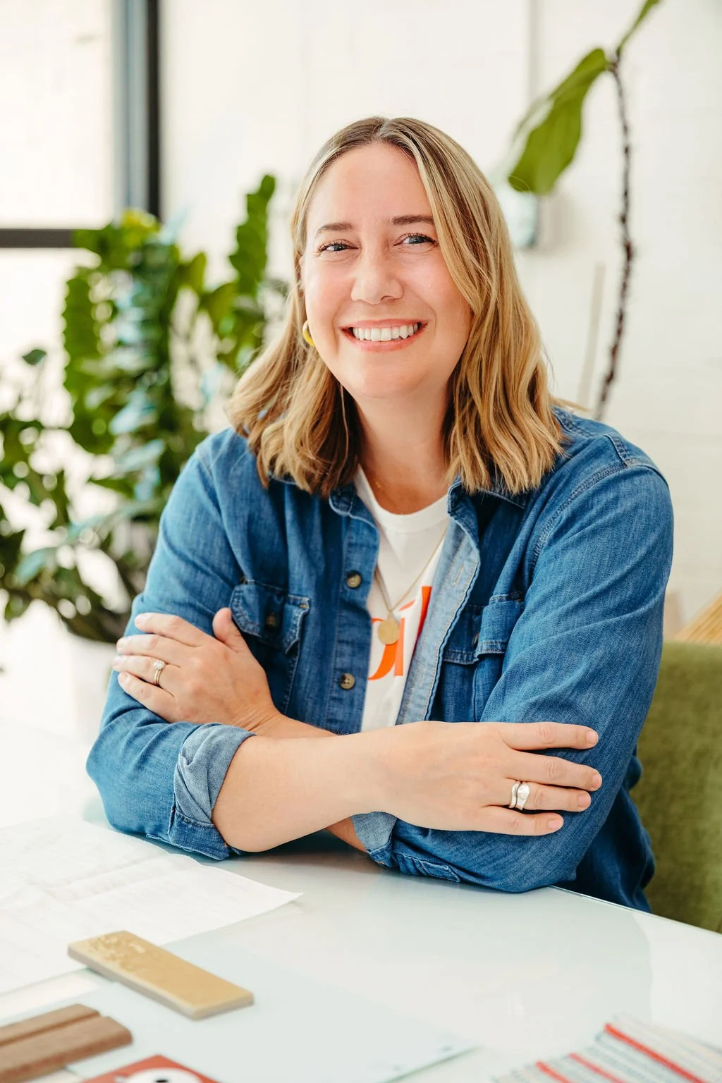 A woman with shoulder-length blonde hair smiling with arms crossed, sitting at a white table in an indoor space with large windows and green plants in the background.