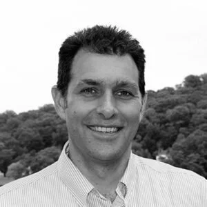 Black and white photo of a man smiling outdoors with trees in the background.