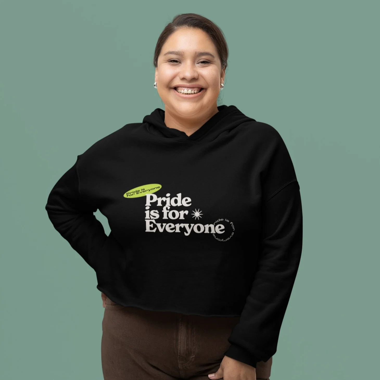 Smiling model posing in front of green wall wearing black "Pride is for Everyone" crop hoodie