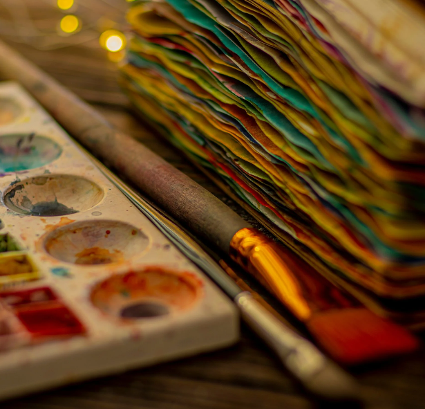 A painting pallet is on the left, paintbrushes are in the center, and a pile of paper is on the right.