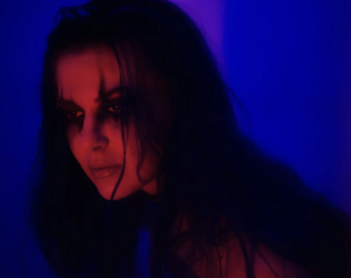 Image of a person whose face is in red lighting, with dark makeup surrounding their eyes. The background of the image is blue.