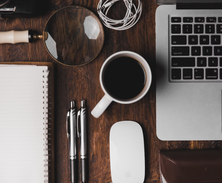 An image of a desk. There are two pens, a coffee mug, a magnifying glass, a computer, and a notebook.