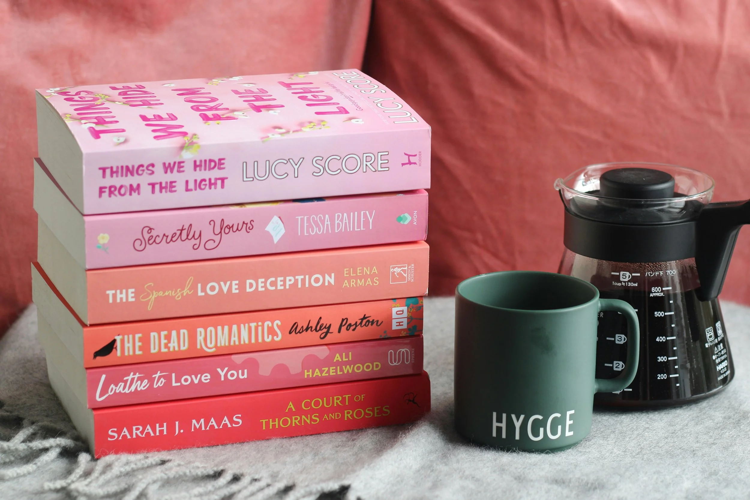 A stack of pink romance books are on the left side. A green mug and a pot of coffee are on the right side. In the background of the photo there are red pillows.
