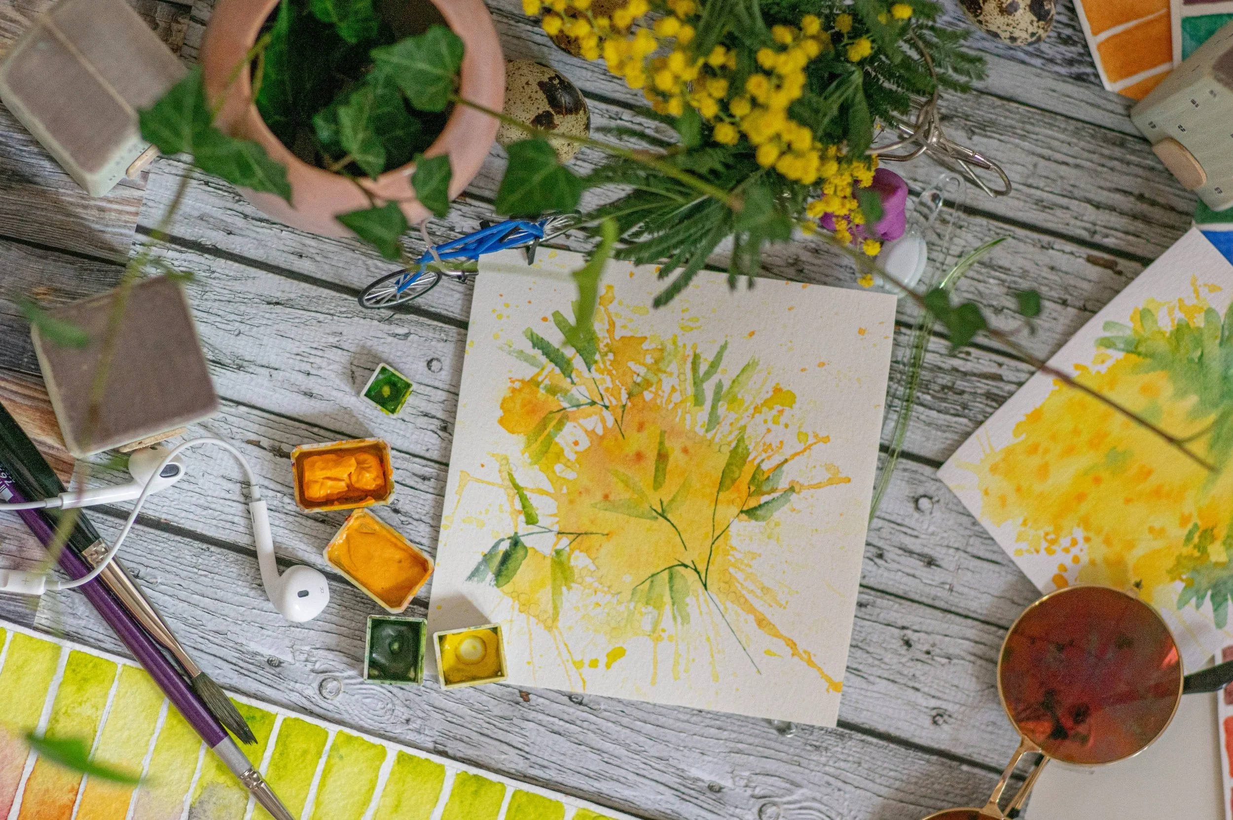 A table covered in painting supplies, plants, and a single headphone.