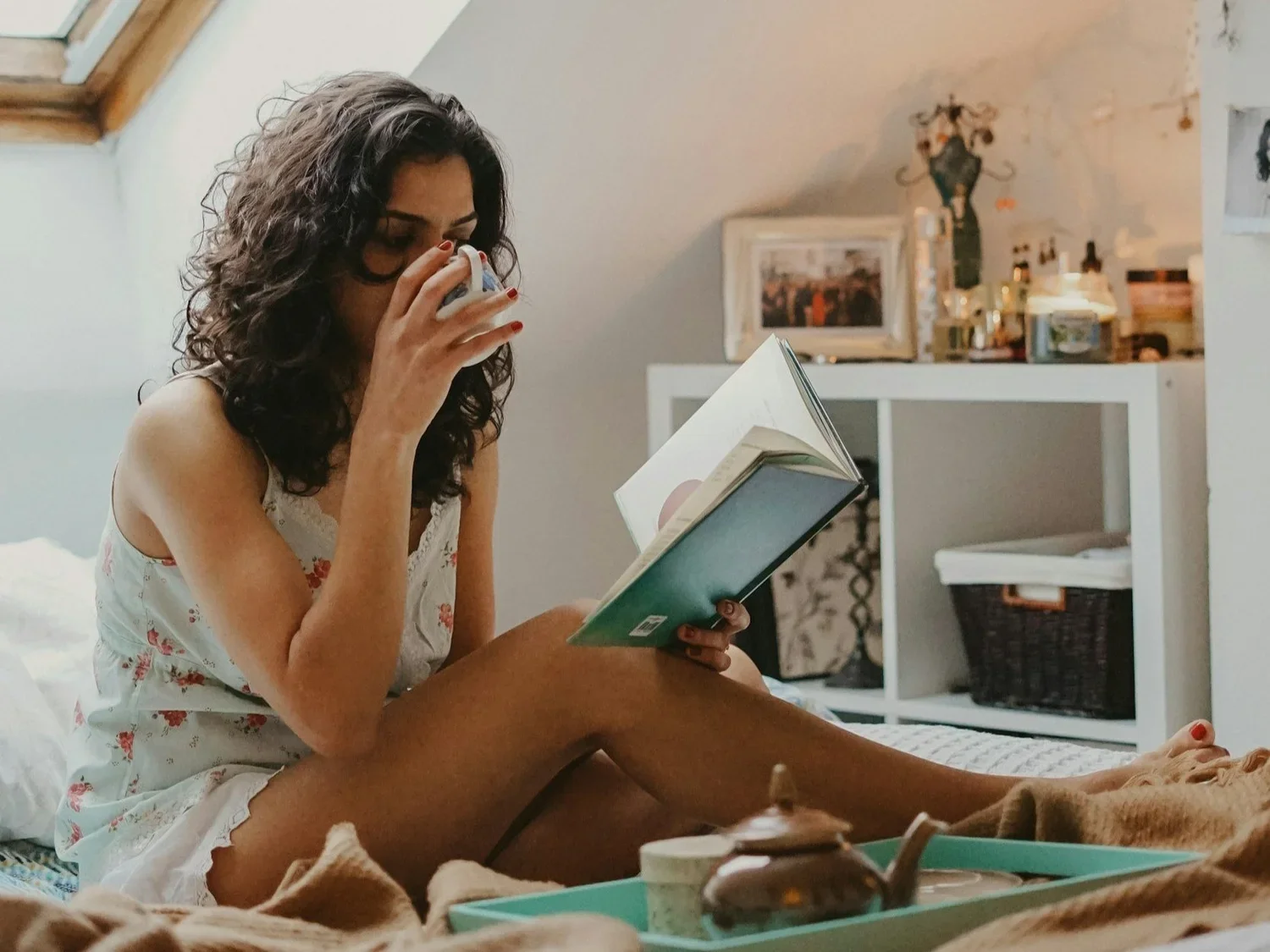A person drinking tea and reading a book in a white walled bedroom. A book shelf is behind them.