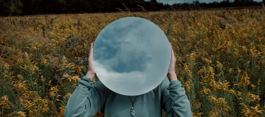 an image of a person in a field with a mirror where their face should be, reflecting clouds