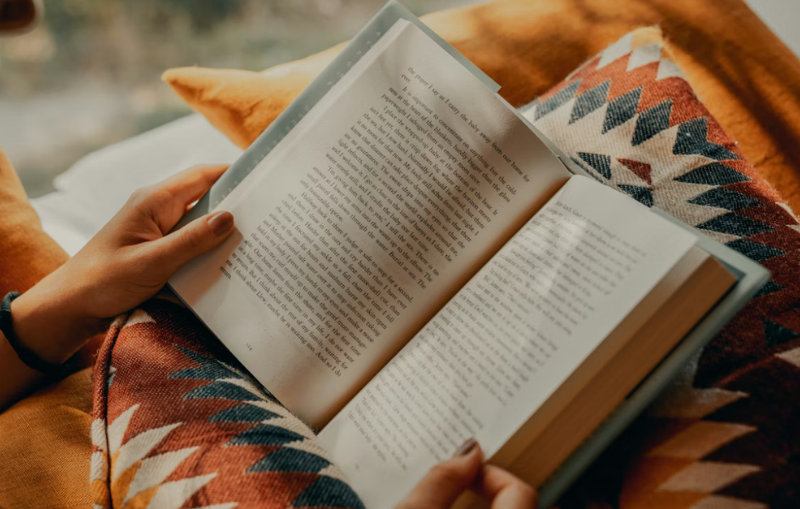 A women is holding open a book, surrounded by orange and cyan pillows
