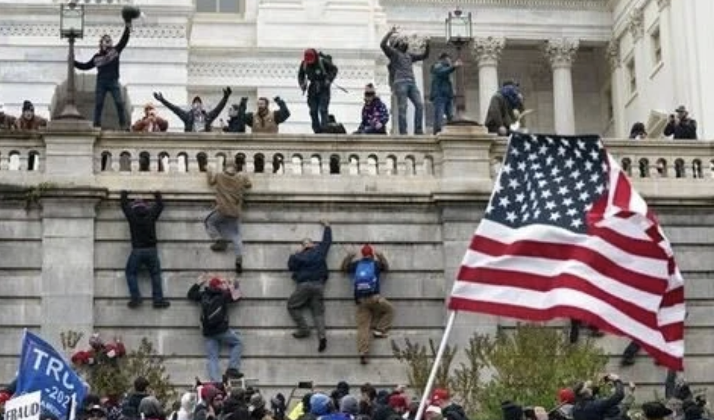 A photo of the January 6th riots with people scaling the wall to enter the White House